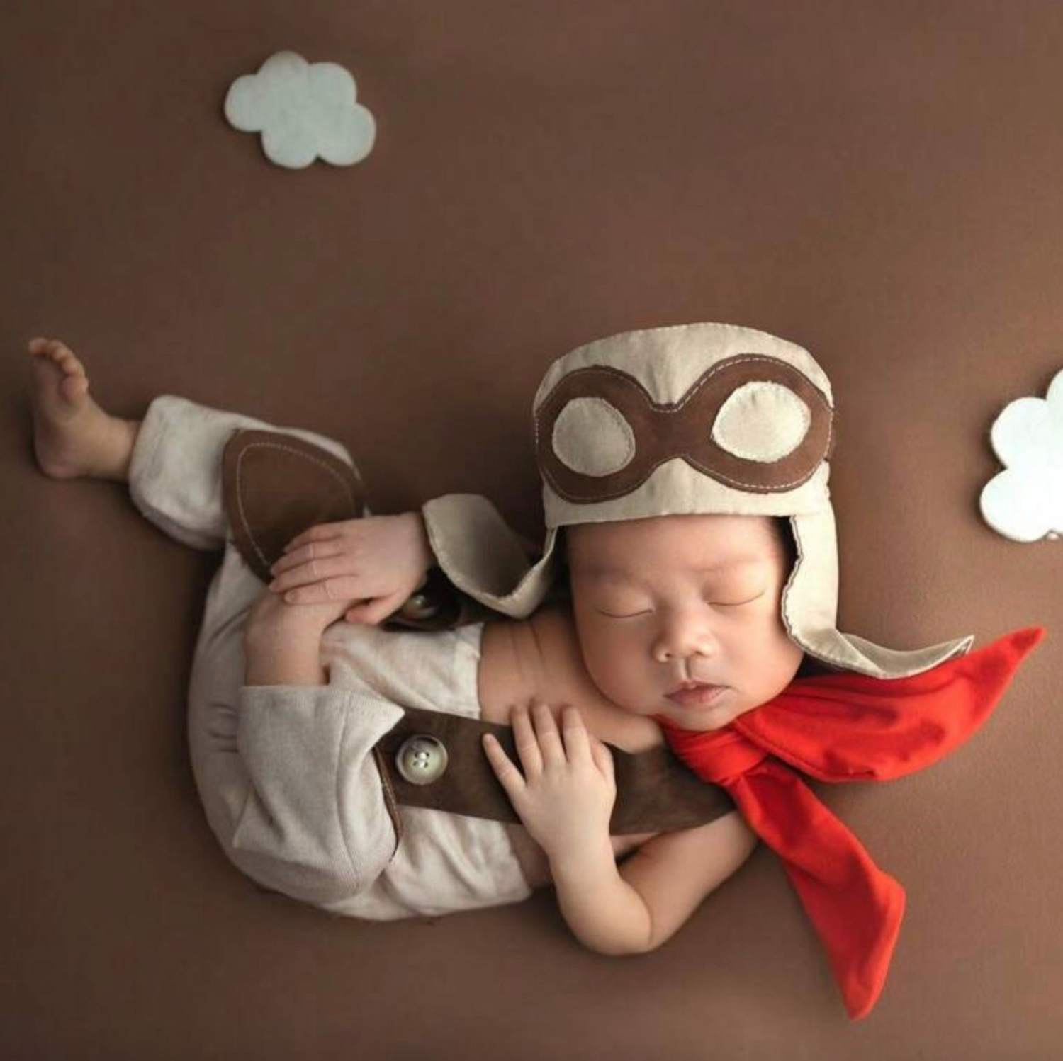 Sleeping baby in gray outfit with red scarf on brown background during winter photoshoot.
