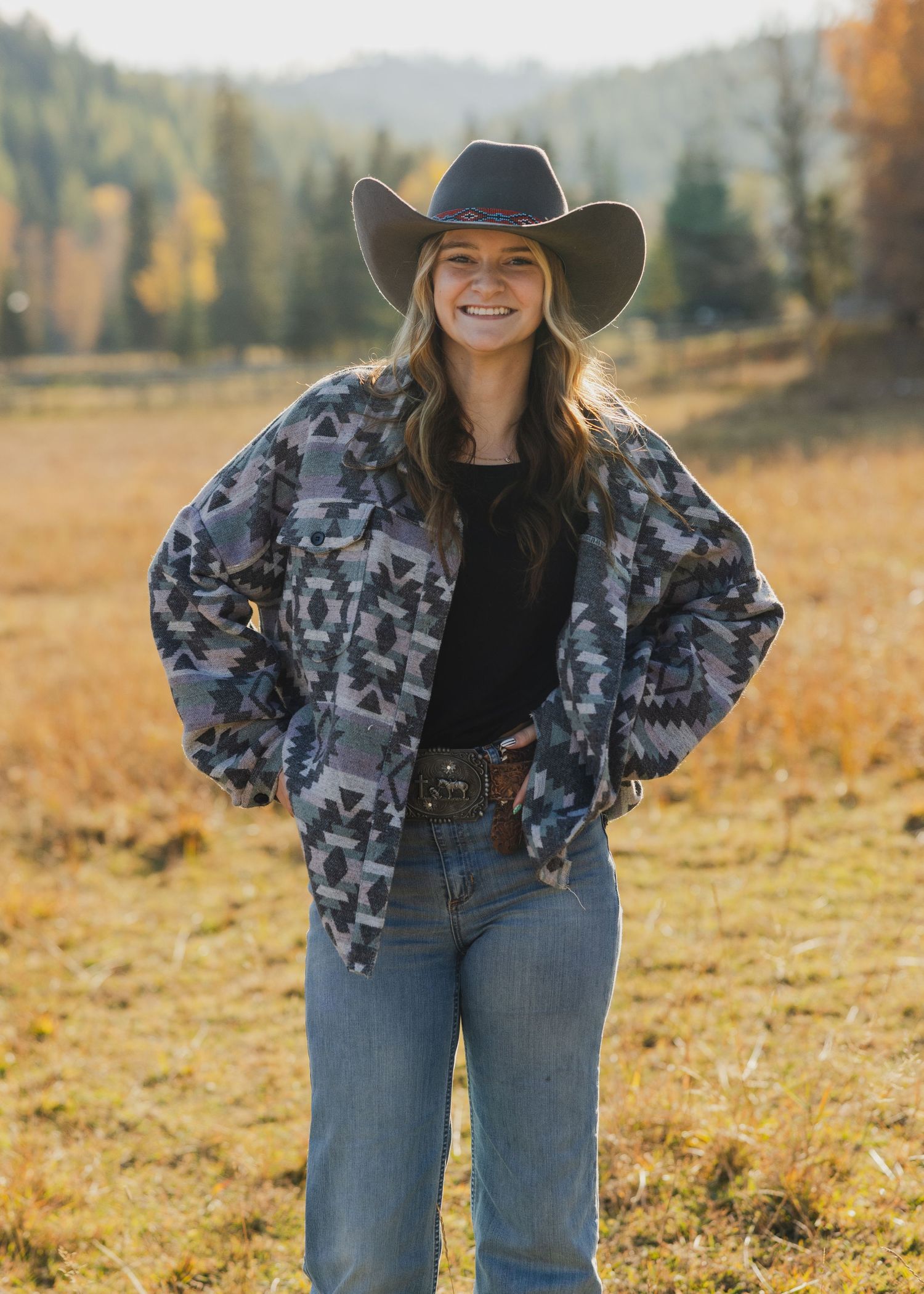 Smiling woman in cowboy hat and Aztec print jacket standing in autumn field.