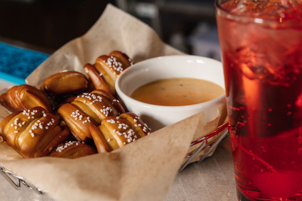 Pretzel bites with dipping sauce and red cocktail served in a wire basket with paper liner.