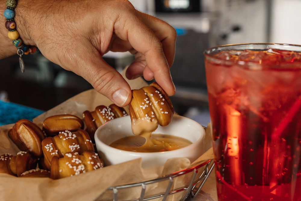 Hand dipping a pretzel bite into sauce next to a red cocktail.