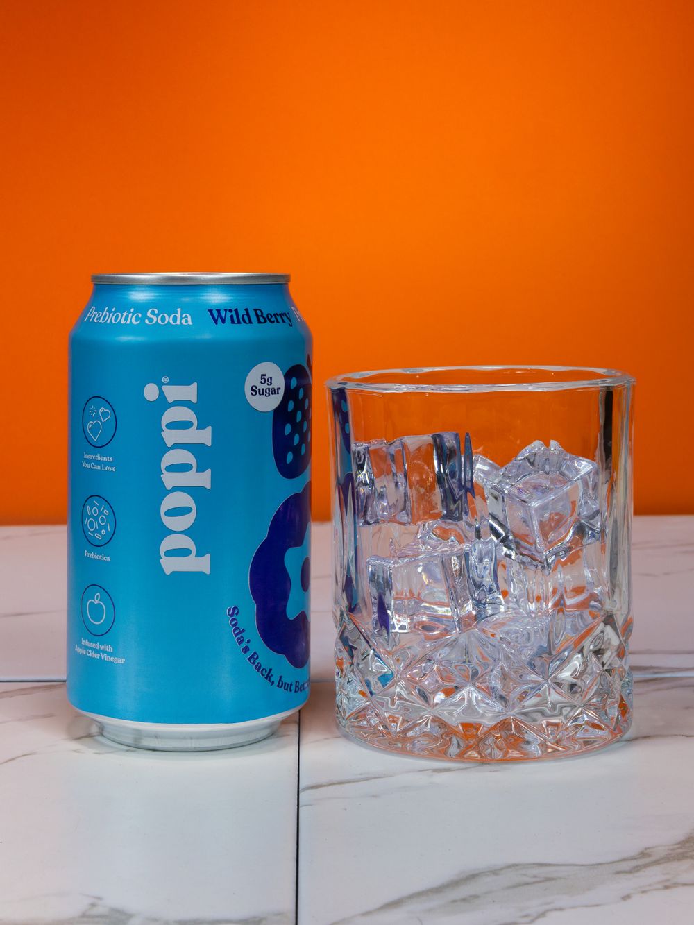Blue Poppi soda can next to crystal glass filled with ice against orange background.