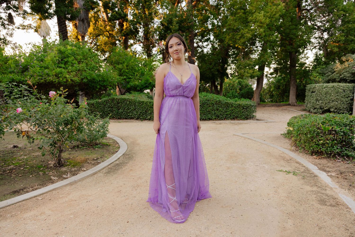 A person stands on a garden path wearing a flowing purple evening gown with a silver belt detail.
