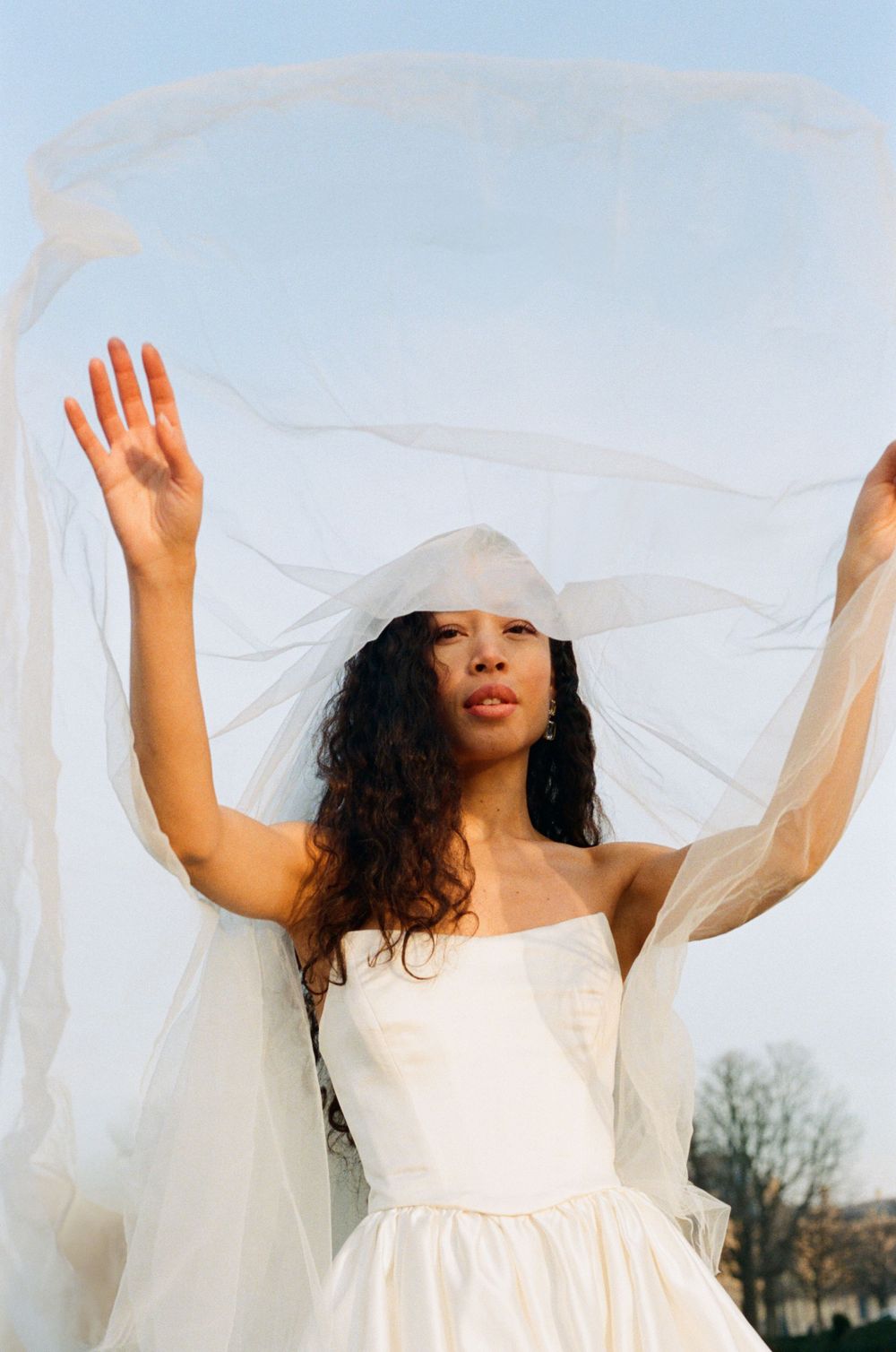 A flowing white wedding dress and veil billow in the breeze as arms are raised gracefully against a soft blue sky.