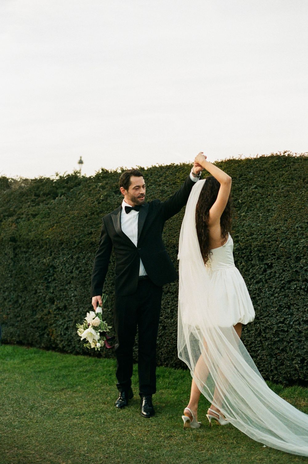 A newlywed couple dances together on a lawn beside a tall green hedge, the bride in a flowing white dress with veil.