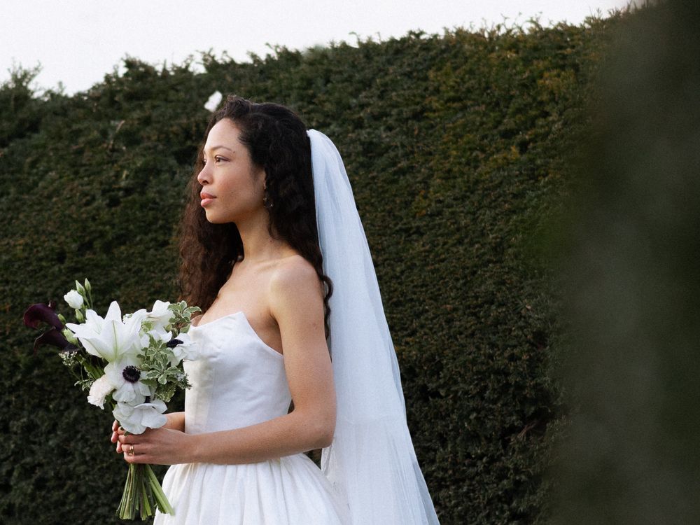 A bride in a white strapless wedding dress holds a white lily bouquet against a hedge background while wearing a long veil.