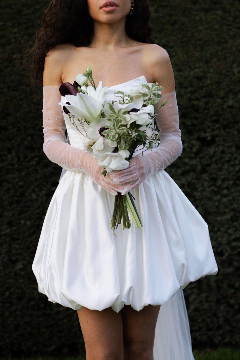 A person wears a white bubble-style wedding dress and sheer gloves while holding a bouquet of white lilies and greenery.