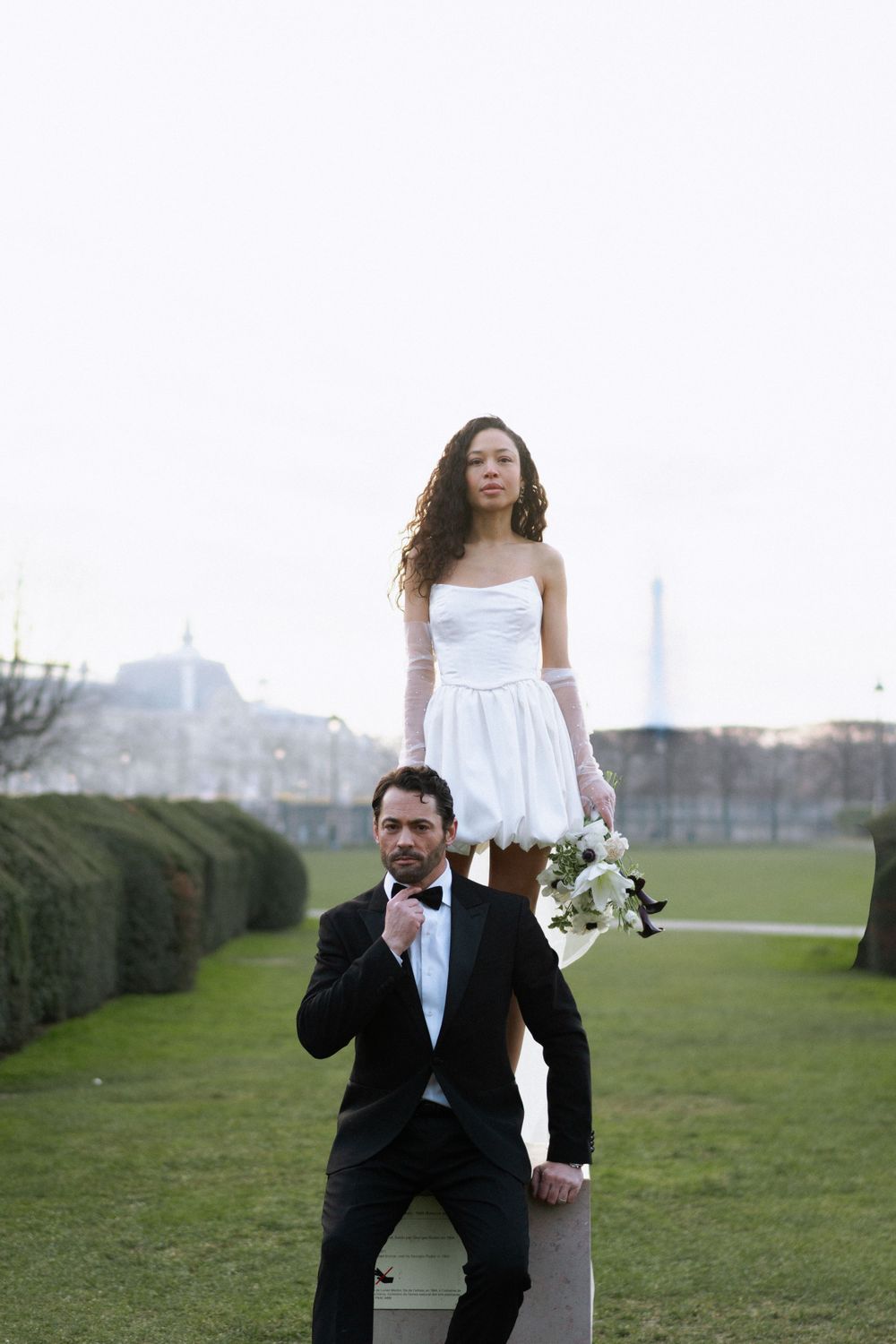Newlywed couple poses creatively in a garden, with the bride standing on elevated ground behind the groom.