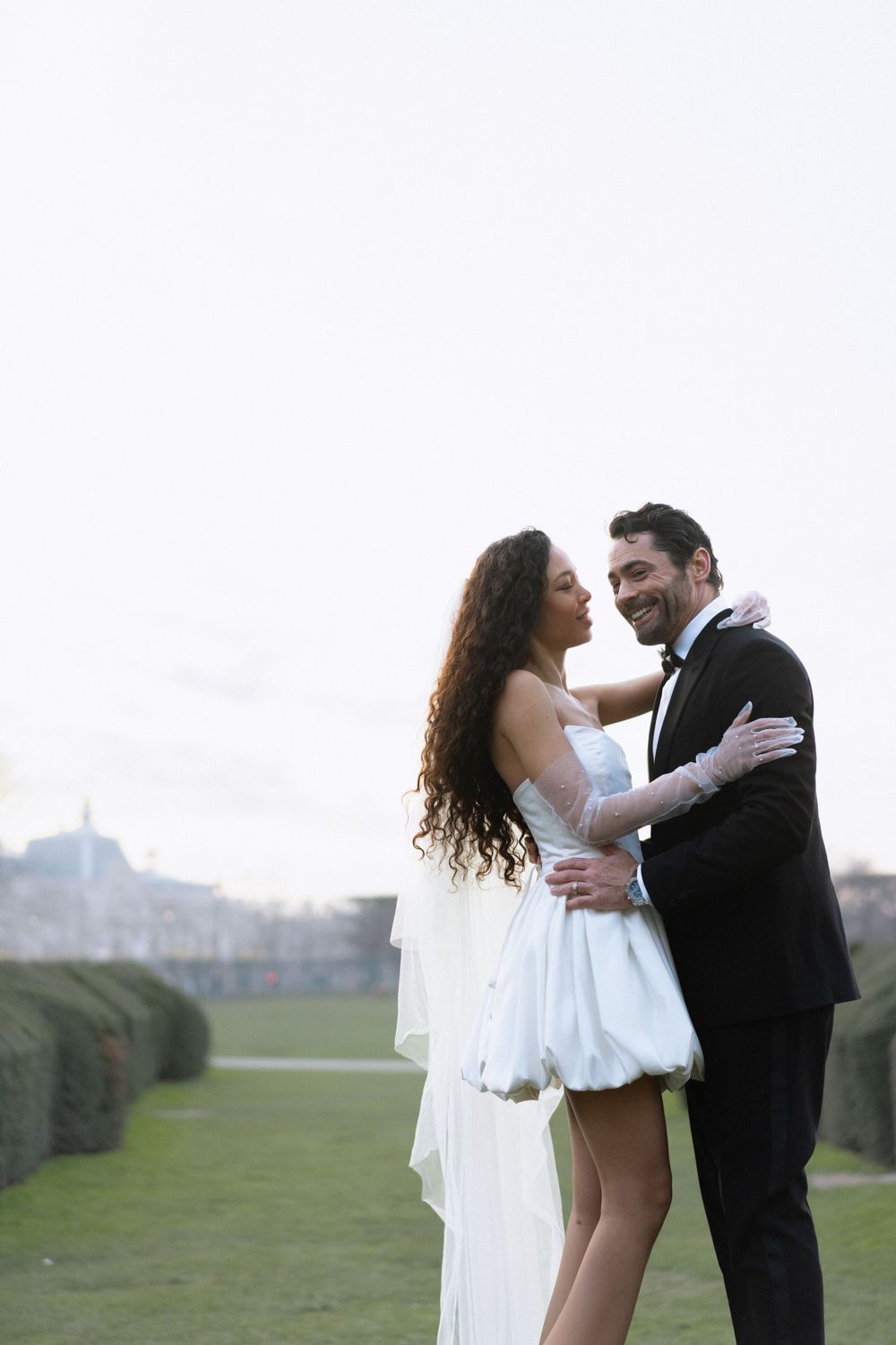 A couple embraces outdoors on their wedding day, with the bride in a short white dress and flowing veil.