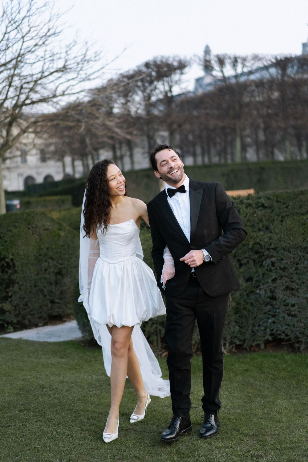 A couple in wedding attire share a joyful moment outdoors in a garden setting with hedges and trees in the background.