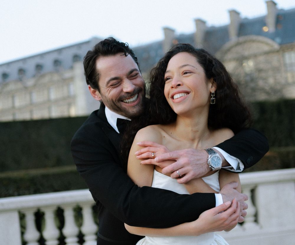A joyful couple embraces and laughs together on a balcony with historic European architecture in the background.