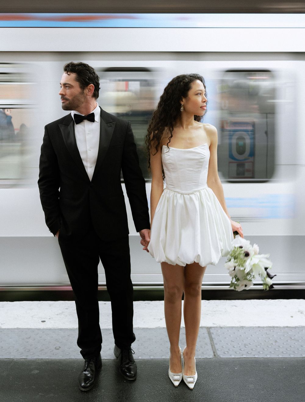 A couple in formal attire, with a white short dress and black tuxedo, hold hands on a subway platform as a train blurs past.