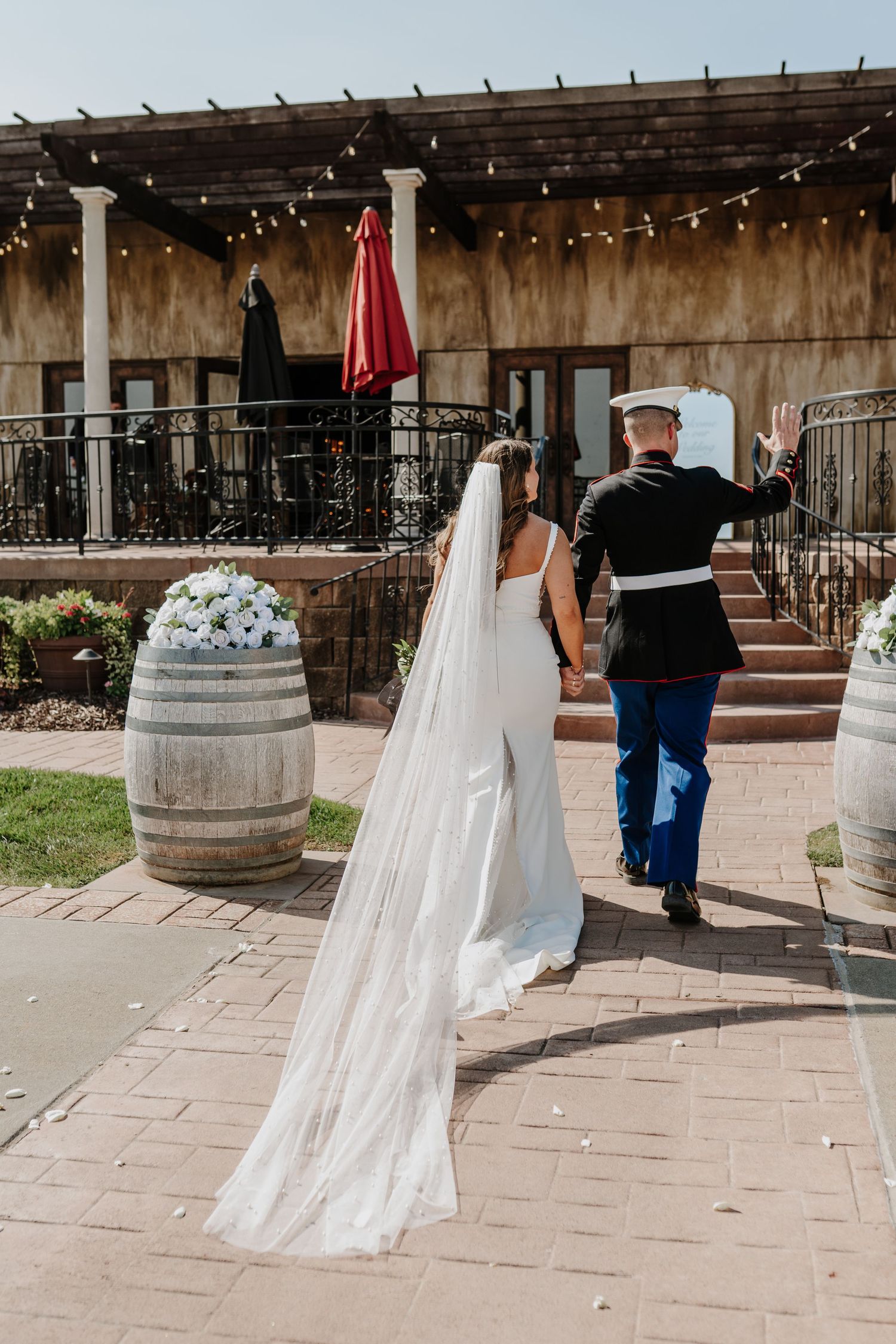A couple walking up stone steps at an outdoor venue with a wine barrel and string lights overhead on their wedding day.