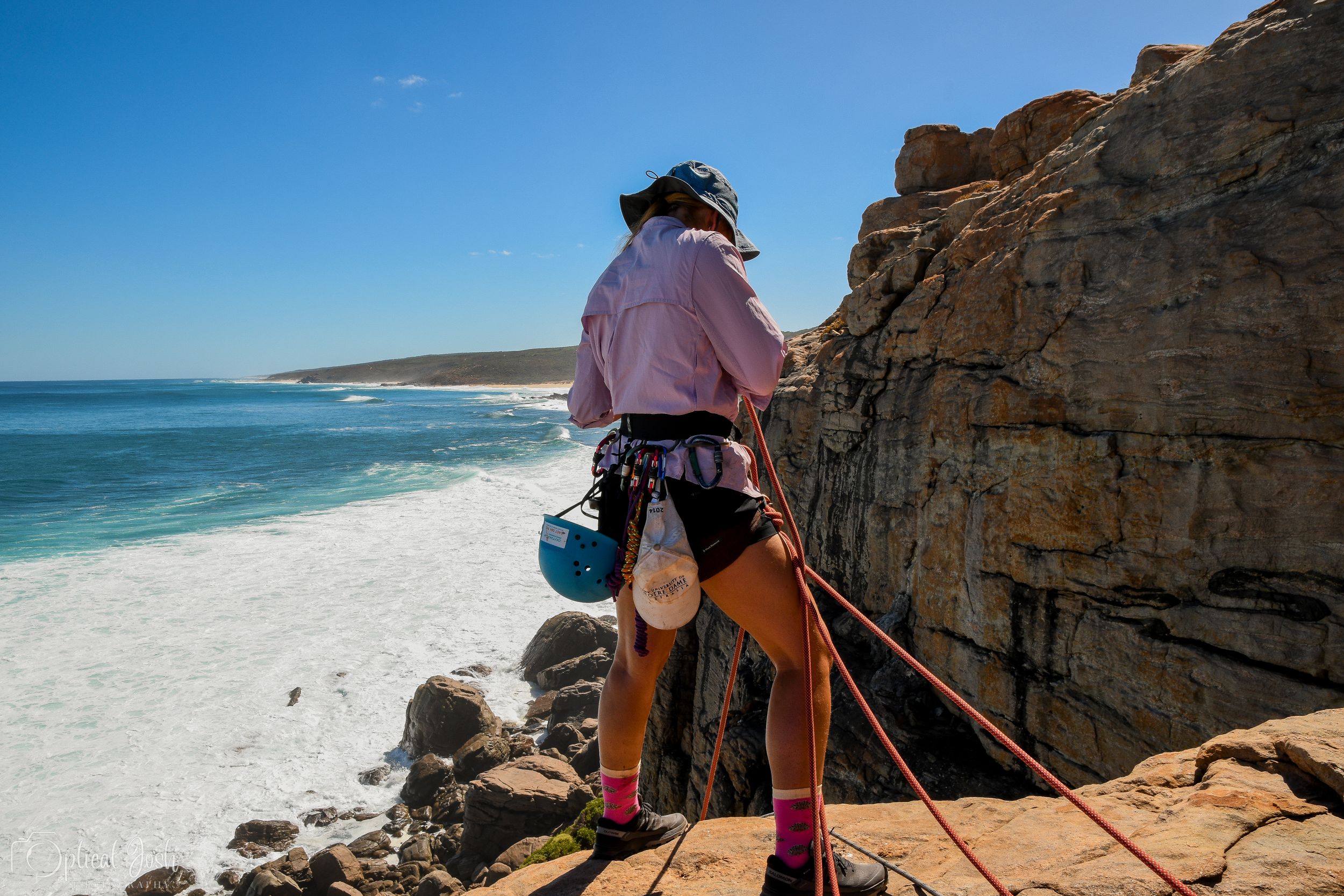 Rock climber in pink shirt and harness stands at edge of coastal cliff overlooking turquoise ocean and sandy beach.