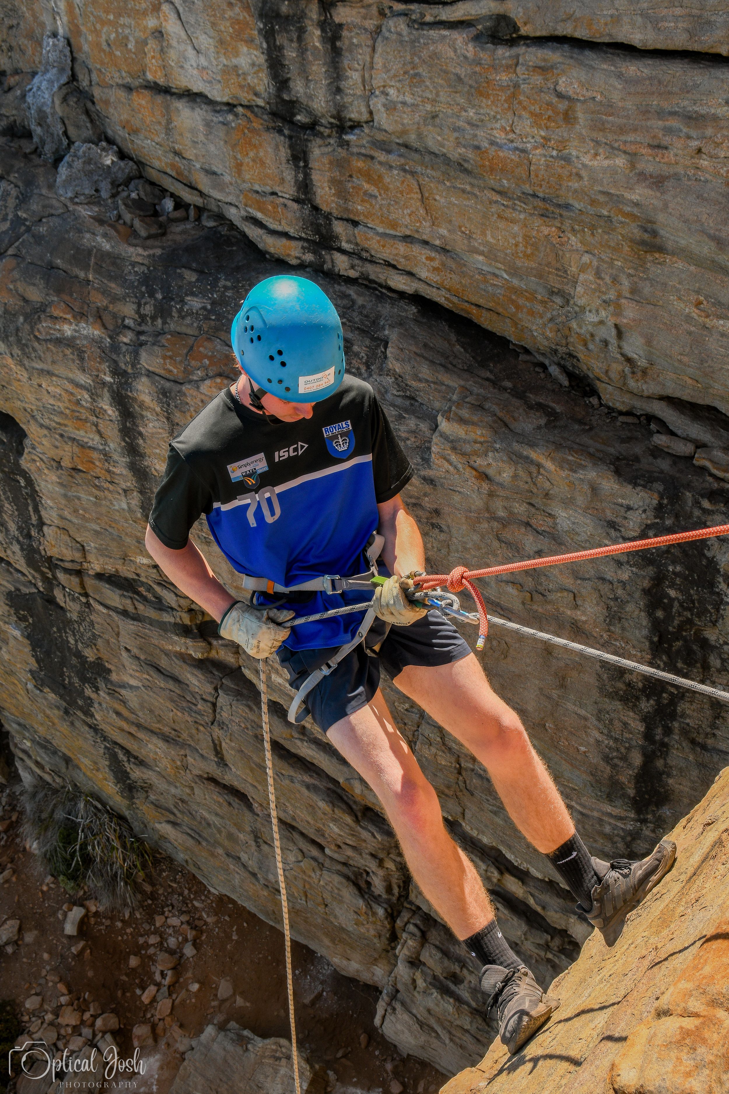 Rock climber in blue helmet and harness rappels down a steep cliff face with orange ropes against rocky terrain.
