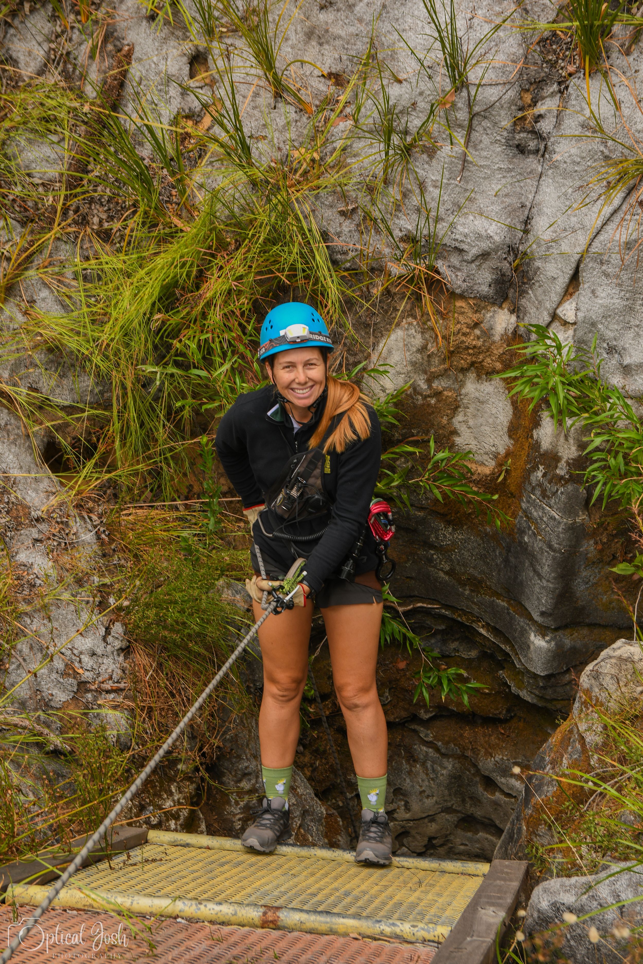 Adventure seeker with safety gear stands on wooden platform while rappelling down mossy rock face in natural setting.