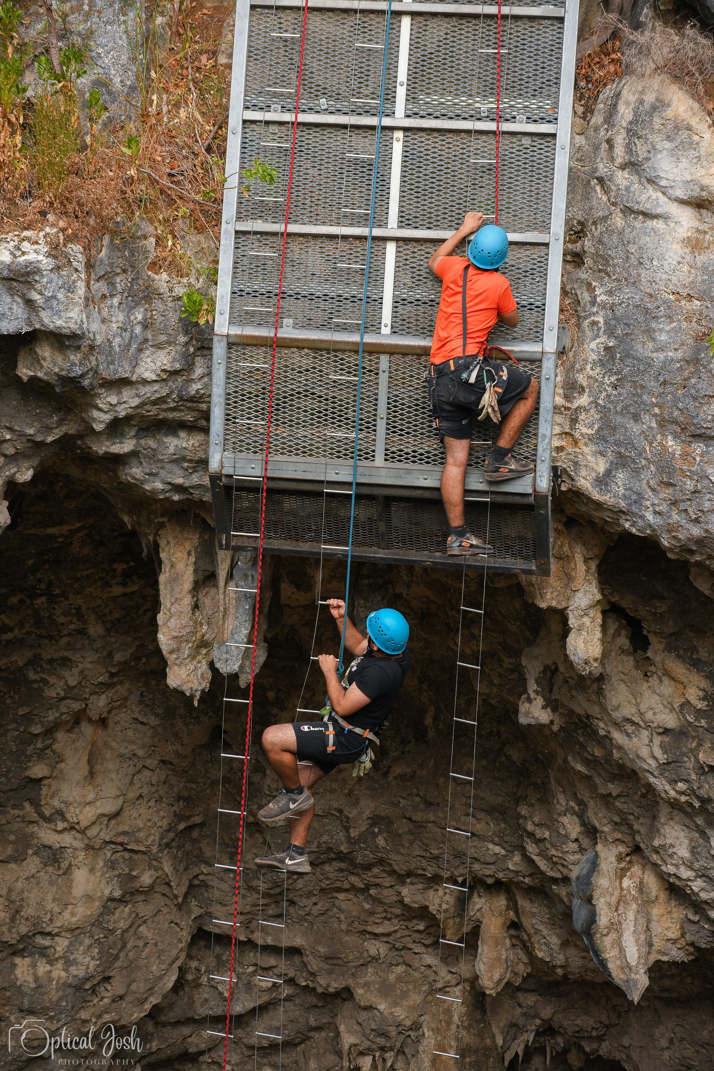 Two climbers in blue helmets and safety gear ascending a via ferrata ladder route on a rocky cliff face.