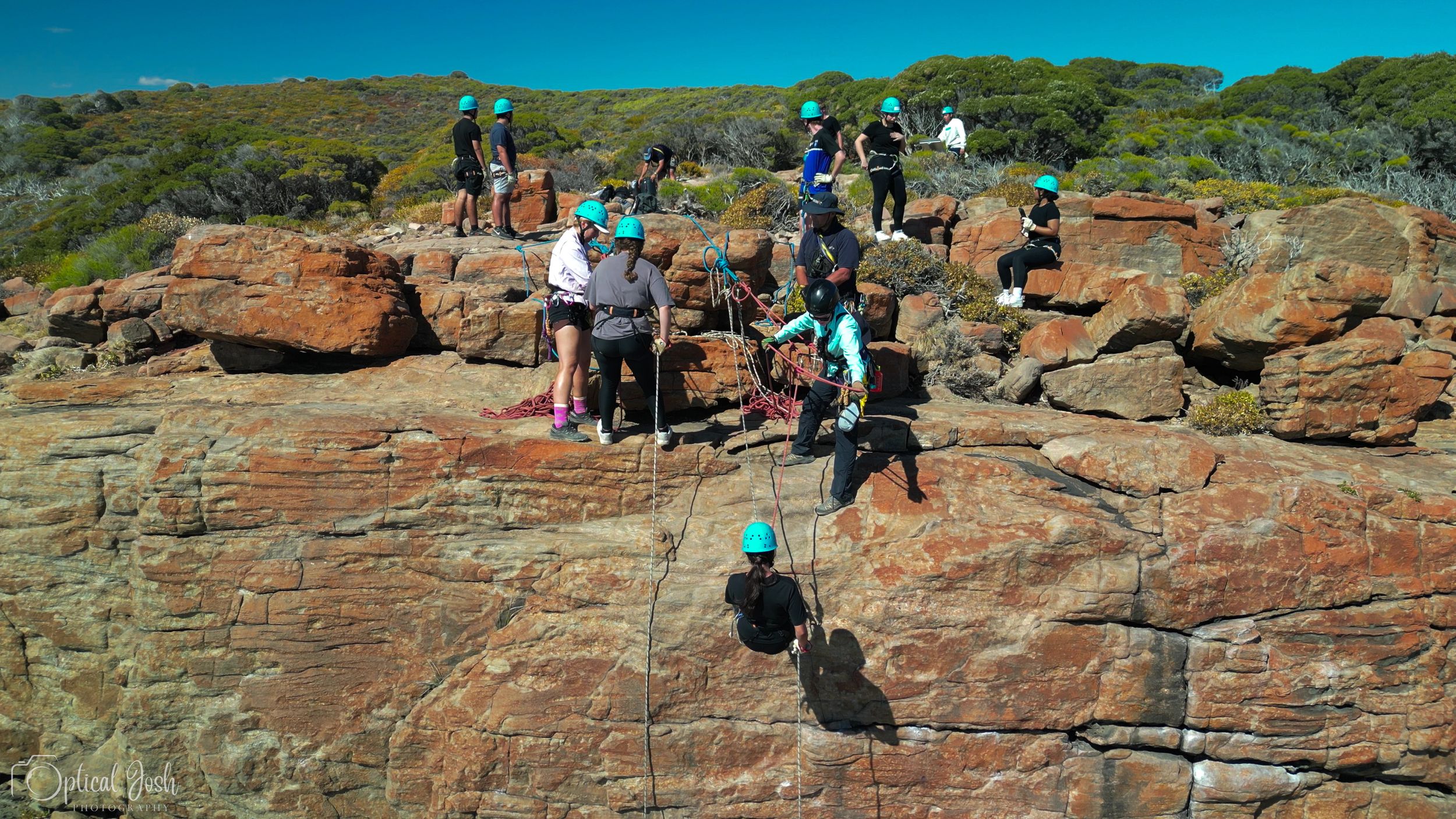 A group of tourists climbing red rock formations along a coastal cliff against bright blue sky.