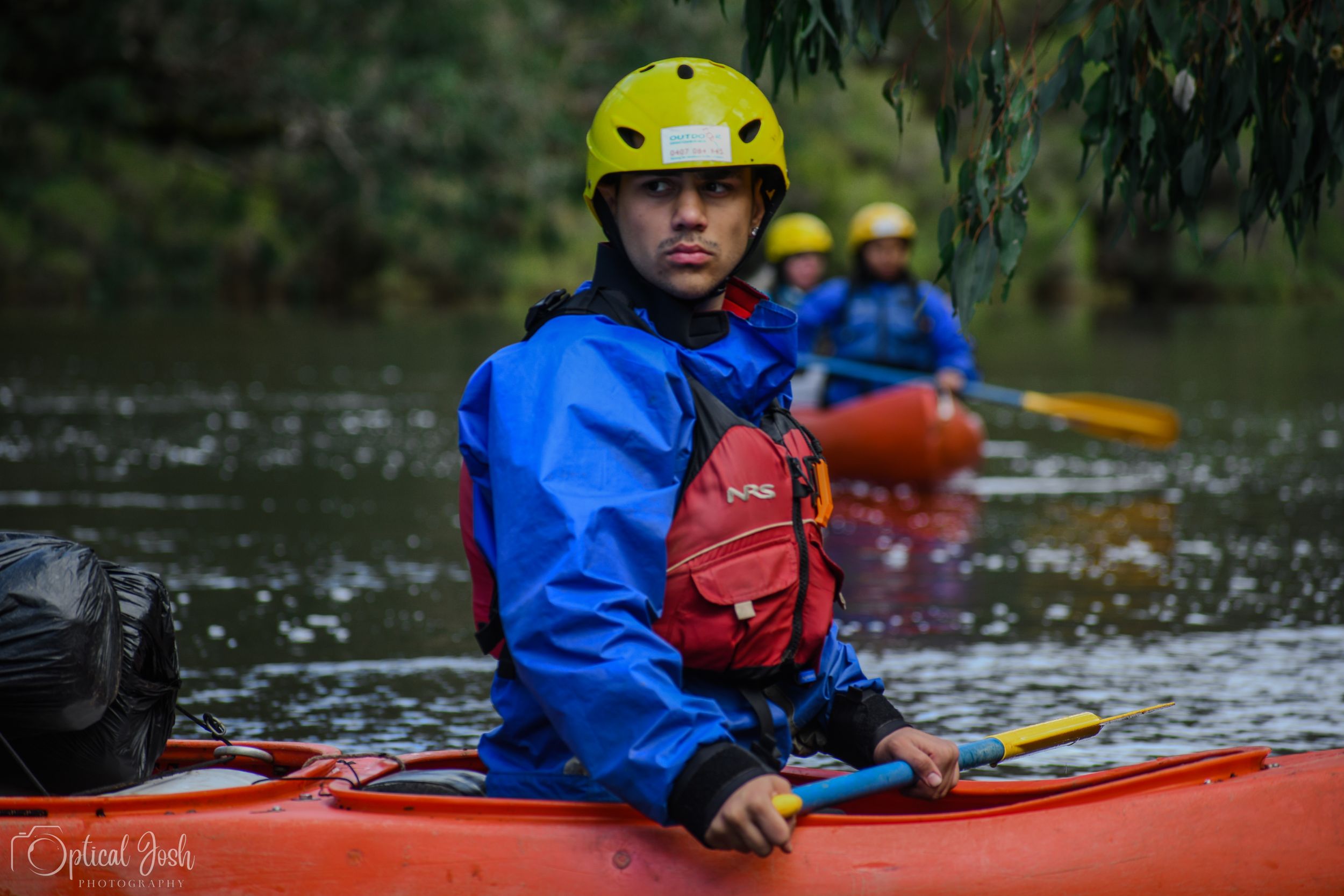 Kayaker in blue waterproof gear and yellow helmet paddling on calm water with another kayaker visible in background.