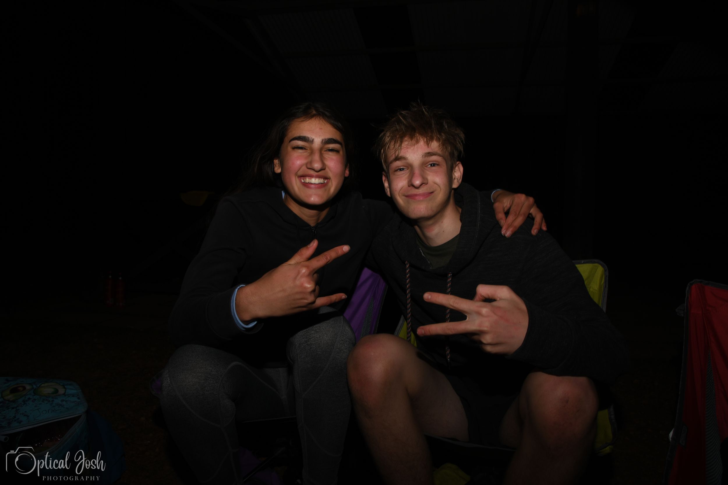 Two friends make peace signs while posing together in a dark setting for a casual photo.