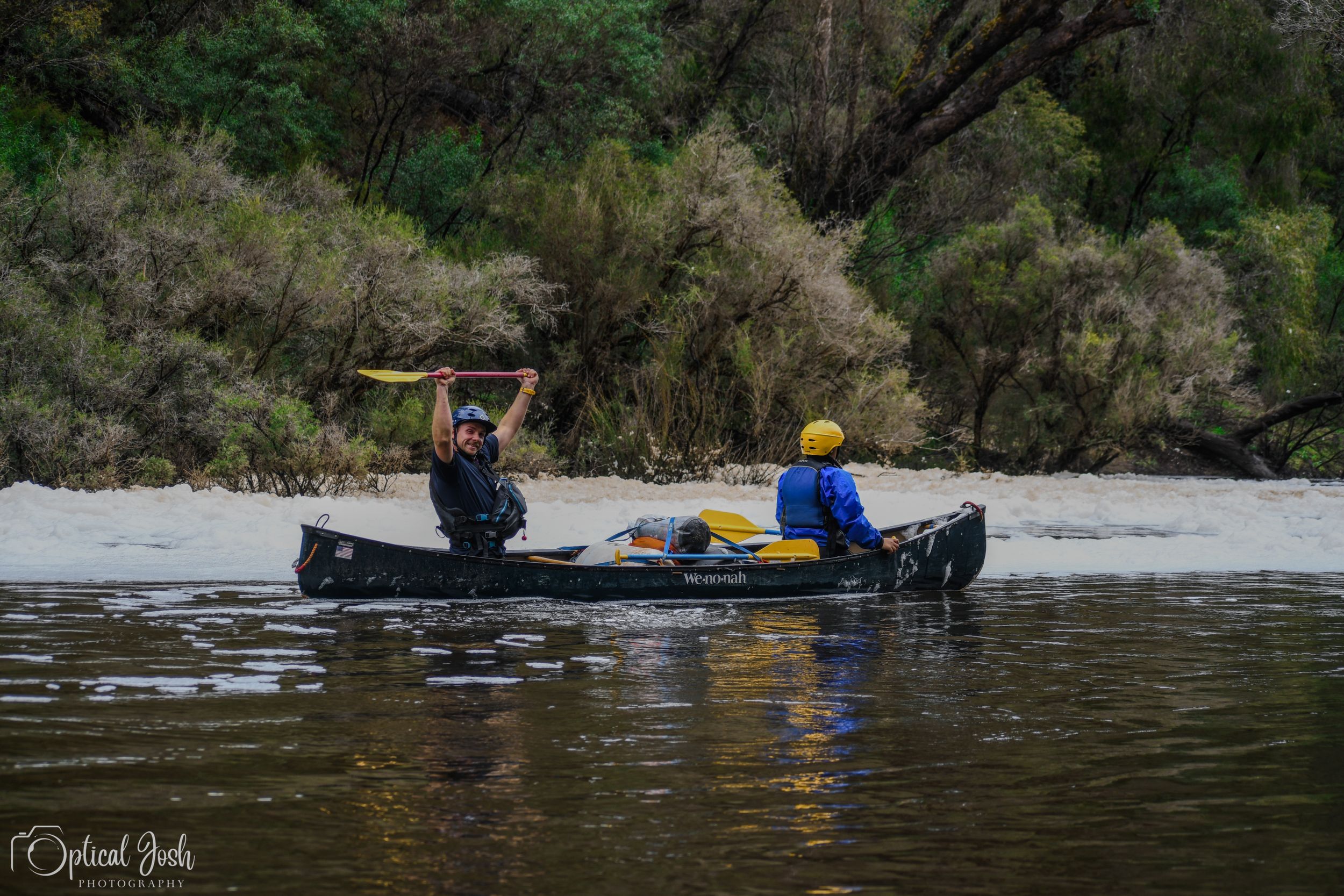Two people paddle a canoe on calm river waters with forested banks in the background.