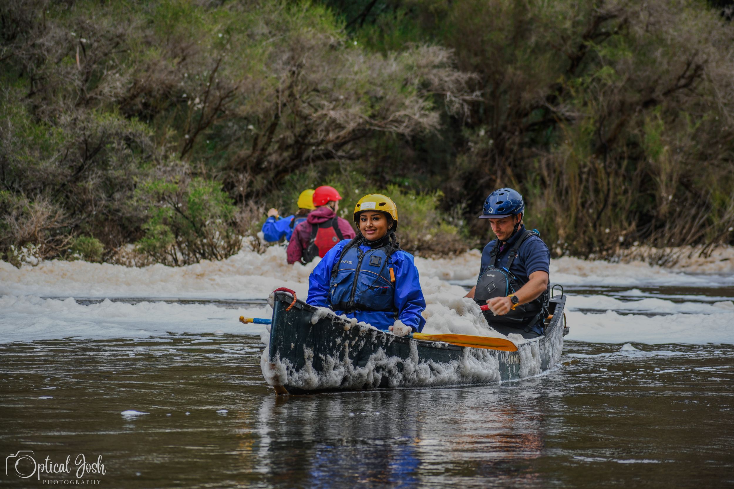 Three people paddling a canoe through white water rapids wearing blue safety gear and yellow helmets.