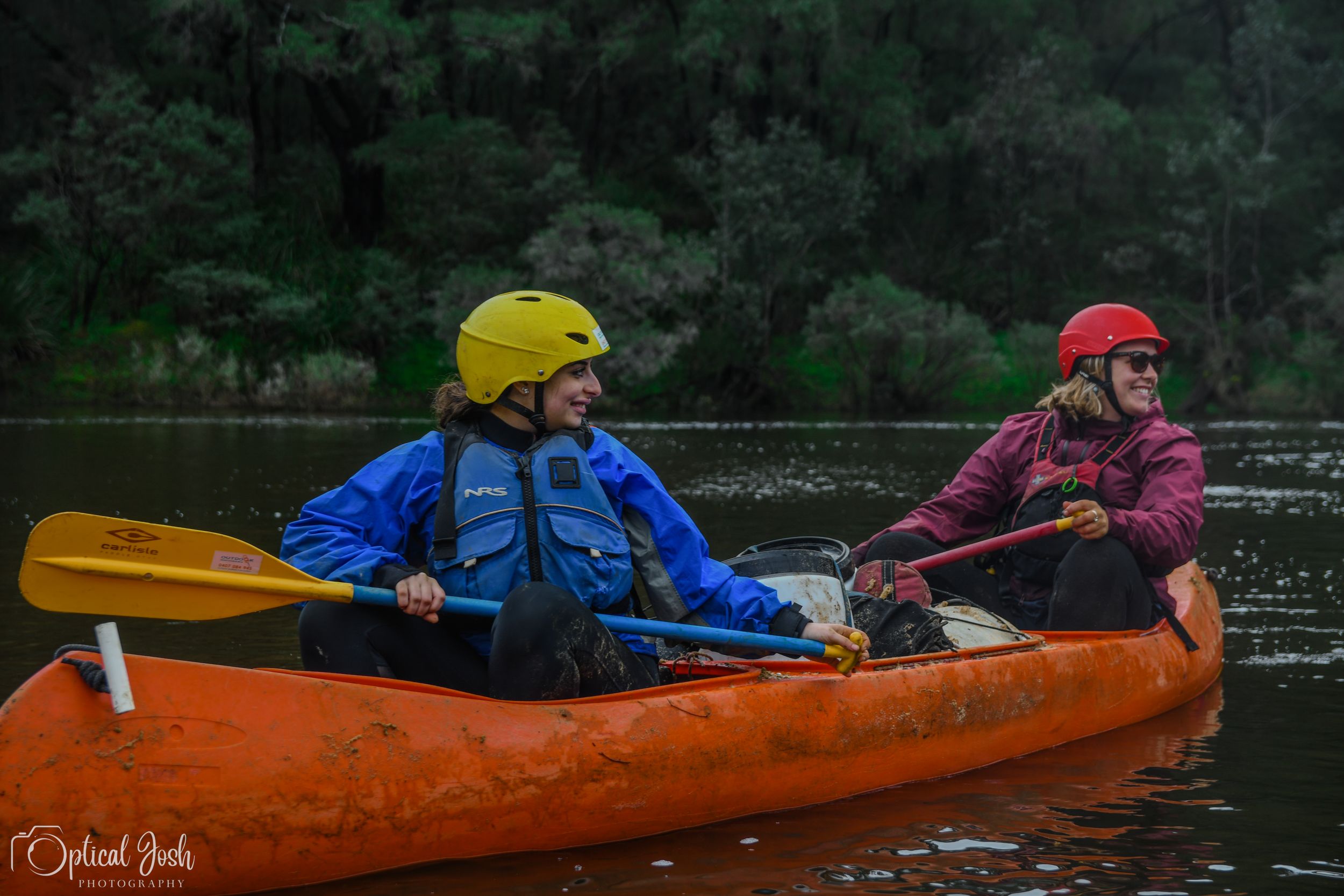 Two kayakers in safety gear paddle an orange tandem kayak across a calm lake on an overcast day.