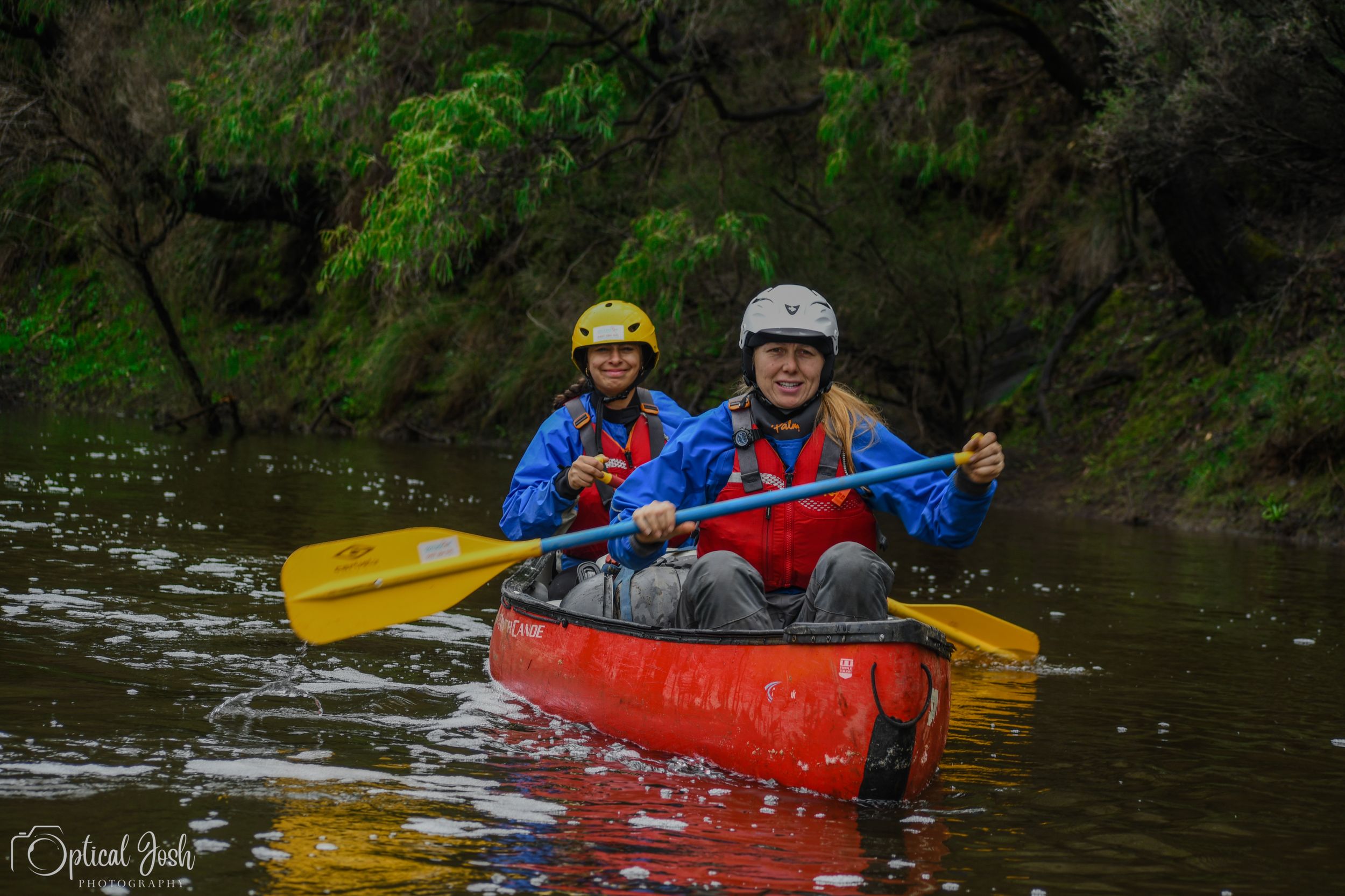 Kayakers paddle a red tandem kayak down a river surrounded by lush green forest.