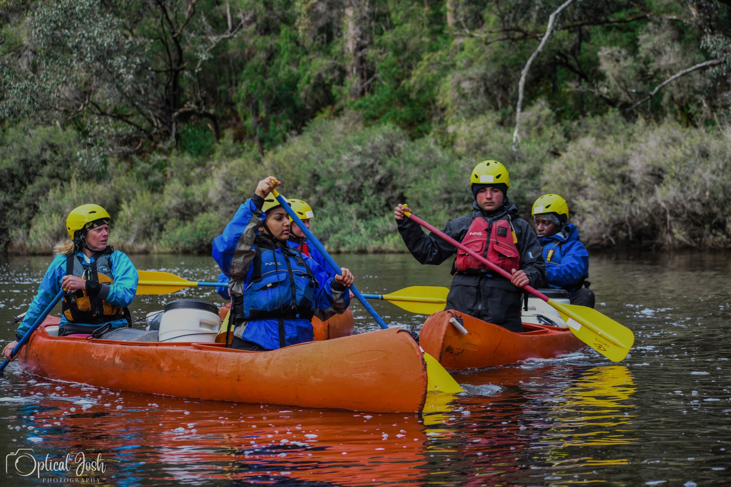A group in orange kayaks paddle through calm waters while wearing blue safety gear and yellow helmets on a river adventure.