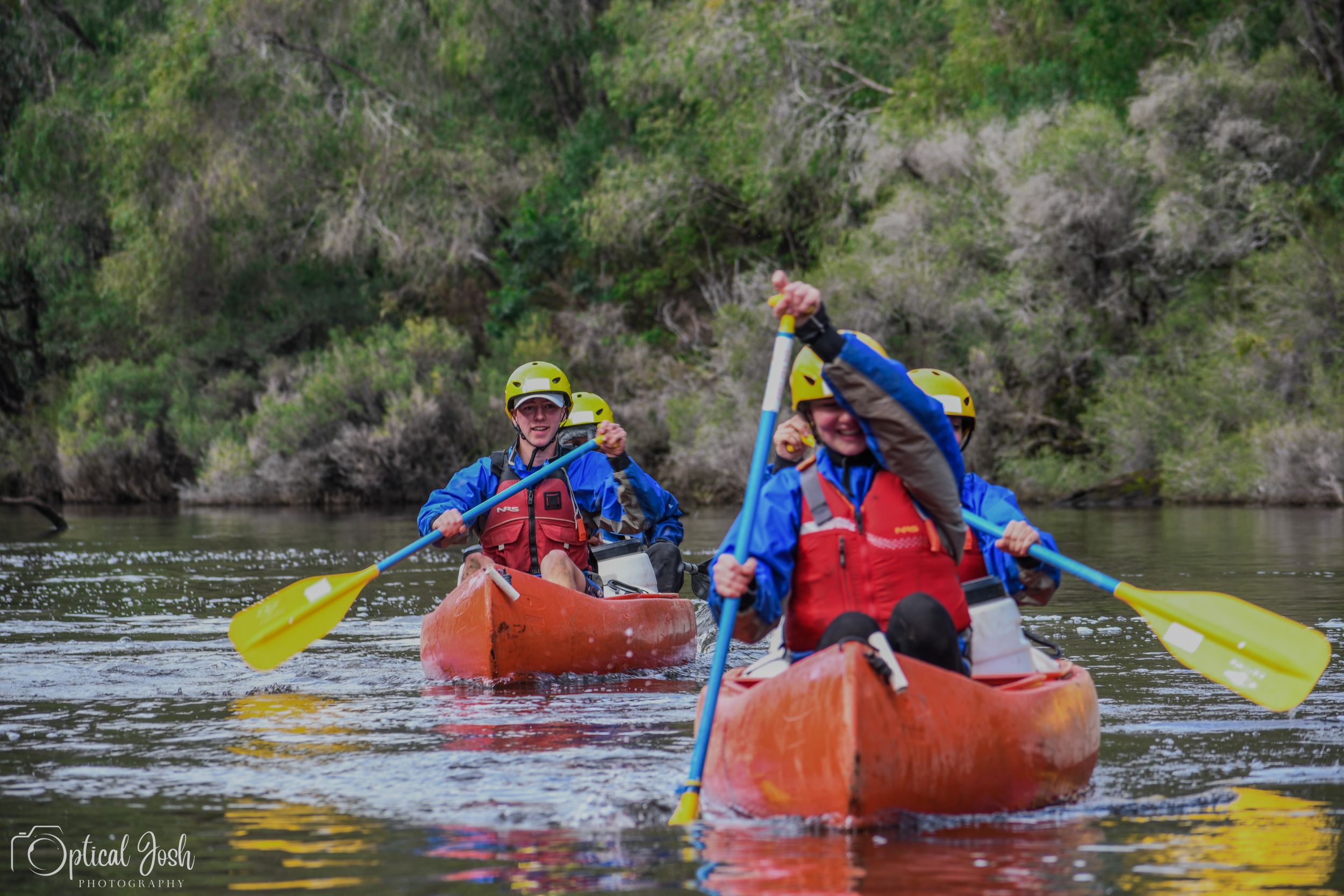 Two paddlers in red kayaks navigate a calm river while wearing blue safety gear and using yellow paddles.