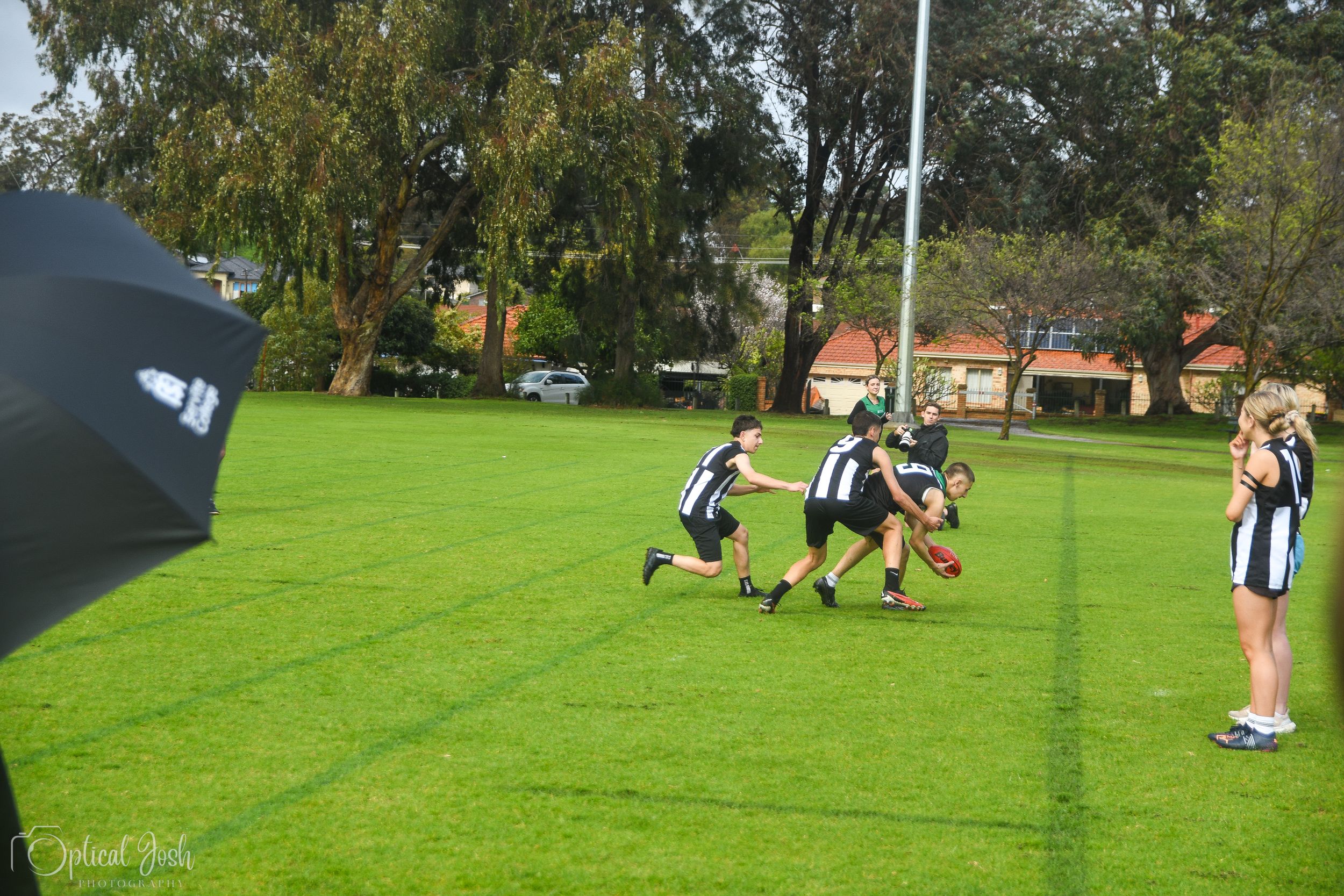 Players compete in an amateur football match on a green grass field with trees in the background.