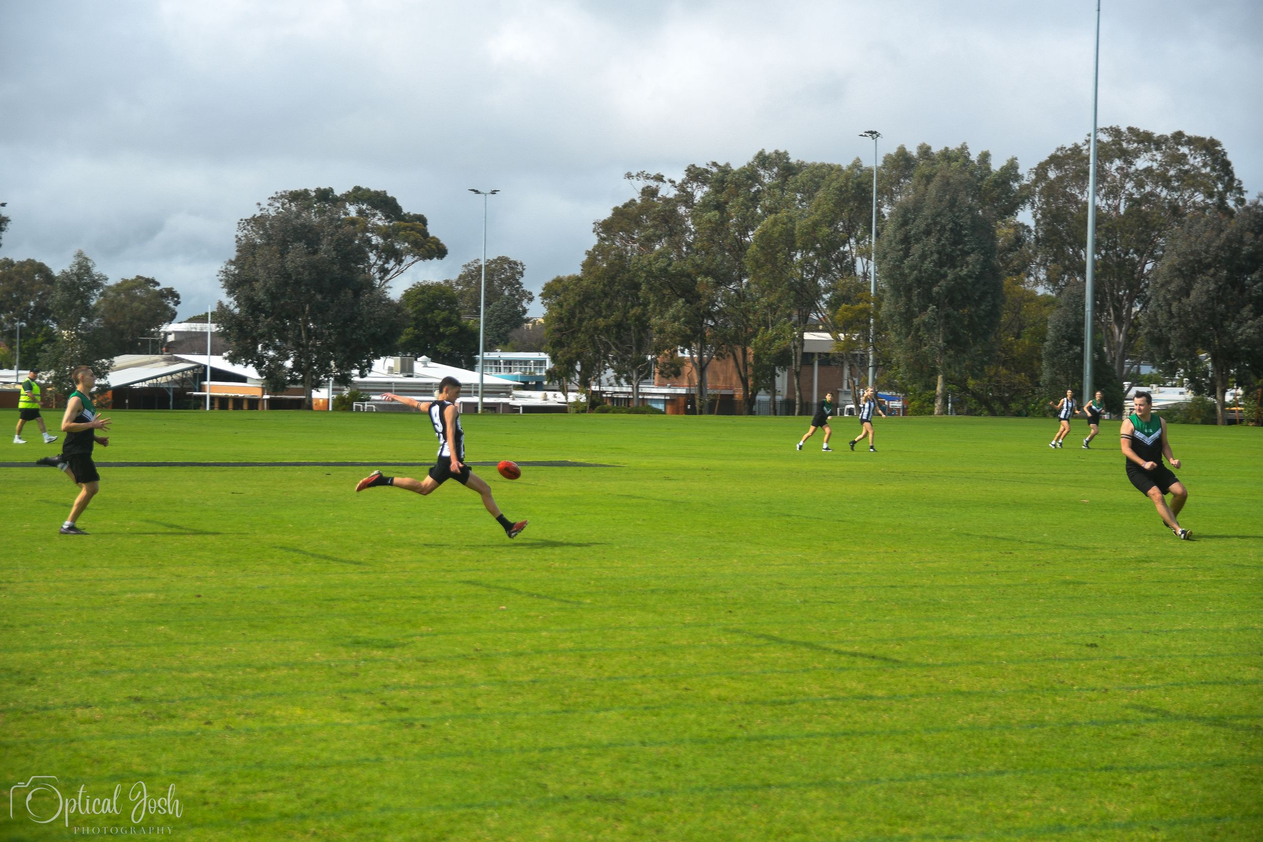 Athletes play on a grass sports field surrounded by trees on an overcast day during a game or practice session.