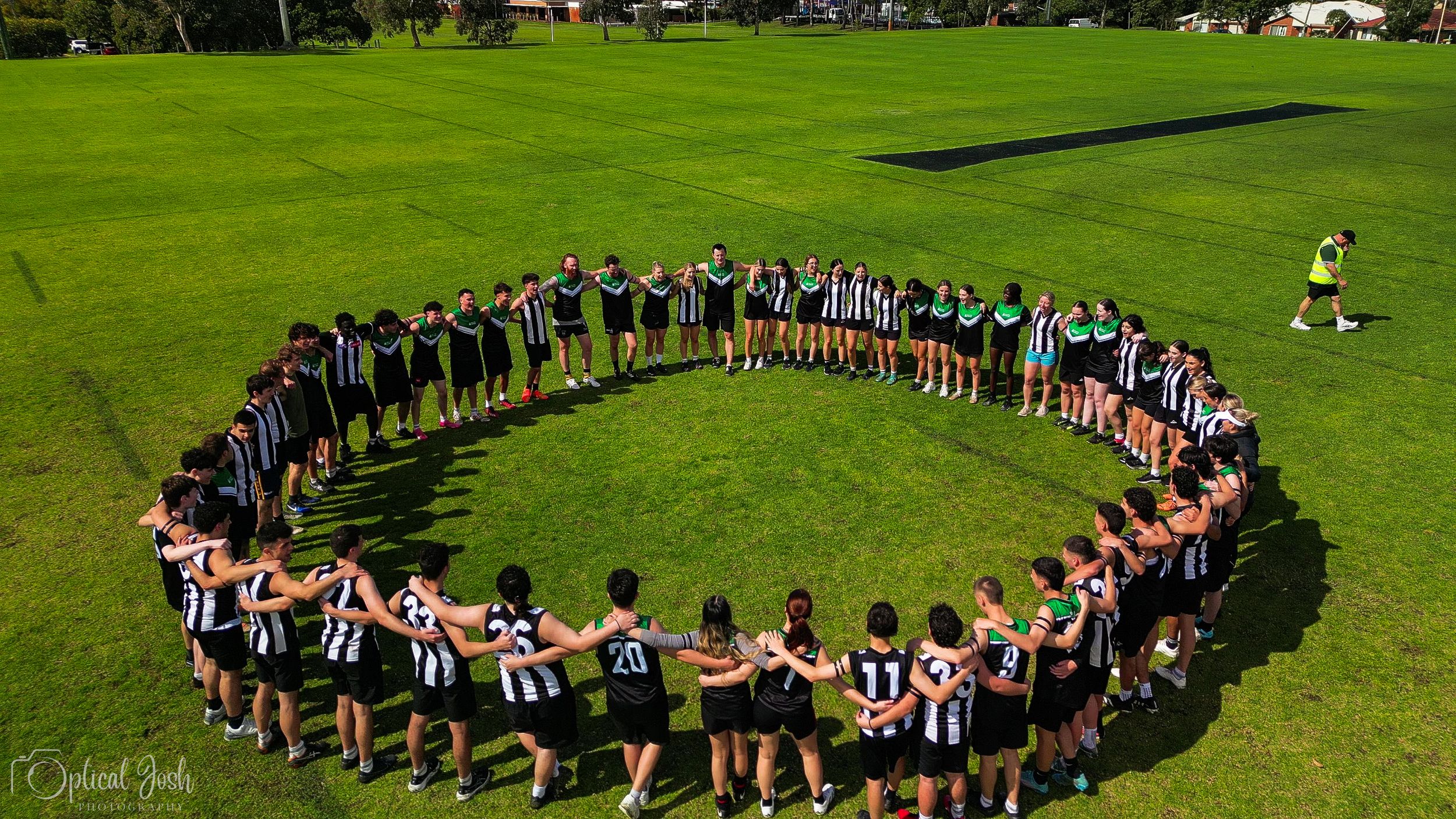 Aerial view of athletes forming a large circle on a bright green sports field during a team gathering event.