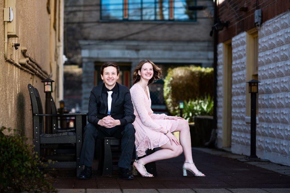 A couple in formal attire sits on stone steps, with the person in pink resting on a higher step than their companion.