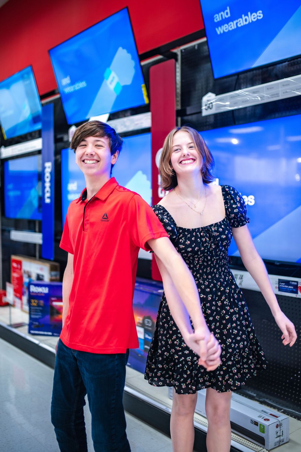 Young couple holds hands while shopping at an electronics store with TV displays visible in the background.