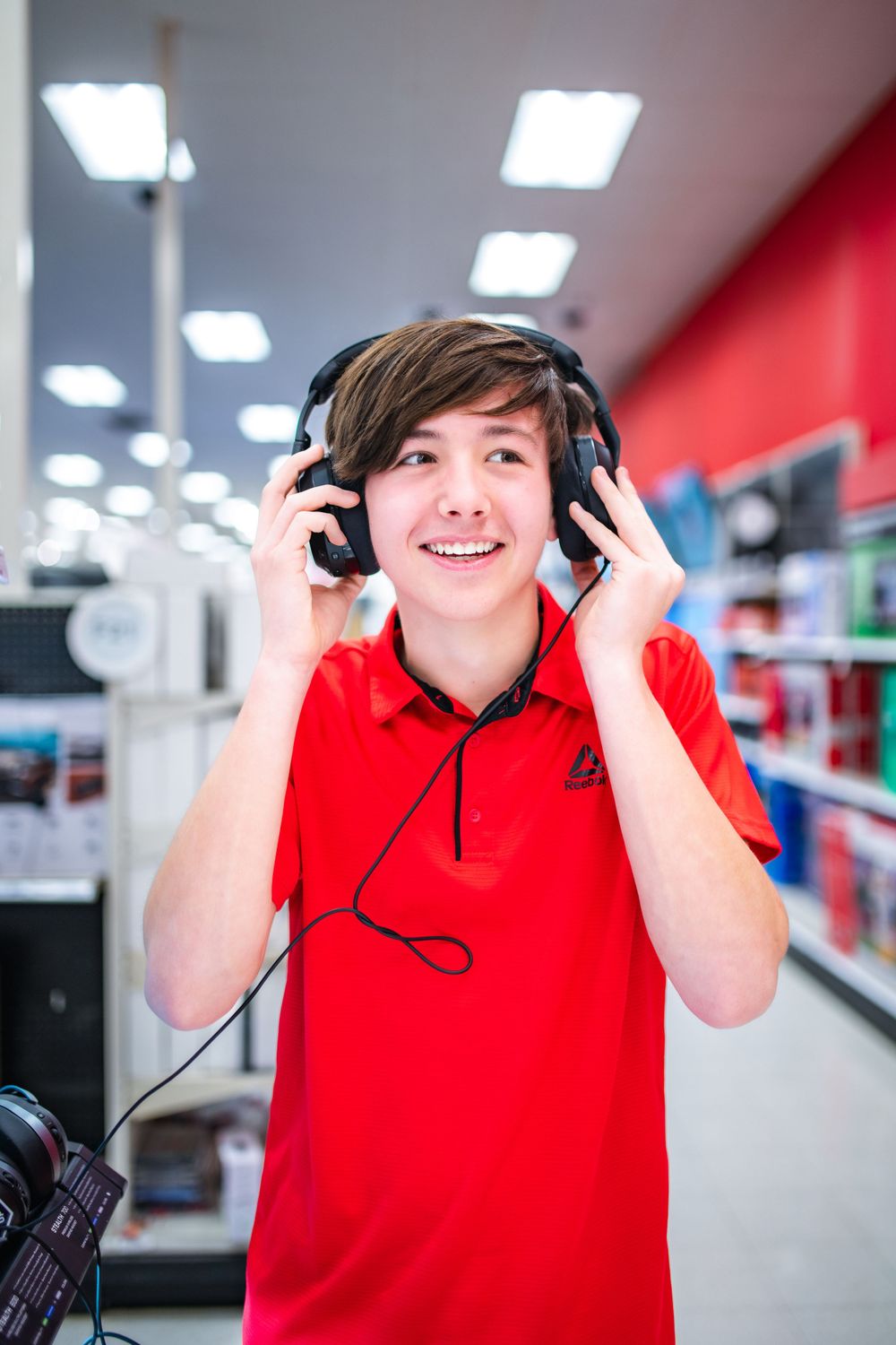 Young retail employee wearing red polo shirt and over-ear headphones smiles while testing audio equipment in store.