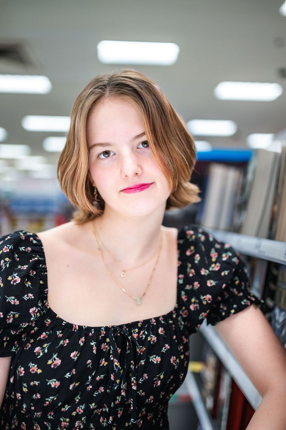 A person wearing a black floral dress and delicate necklace stands in a library or bookstore under fluorescent lighting.