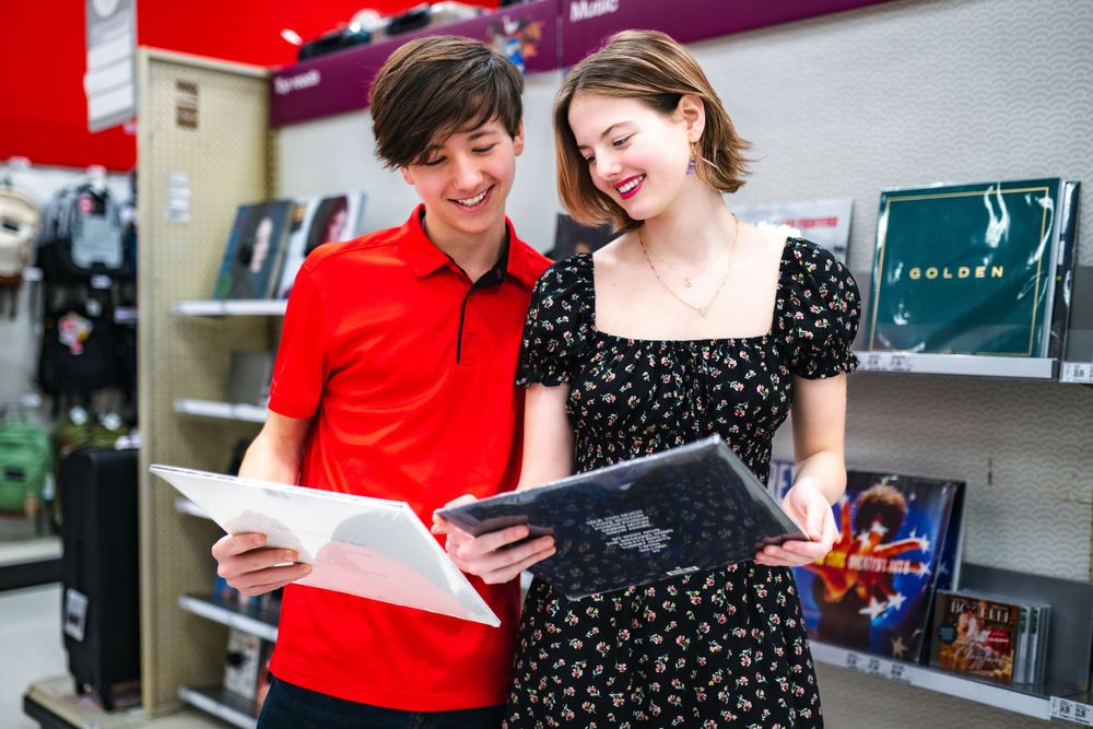 Two friends wearing a red polo shirt and black polka dot dress laugh together while reading documents in a store.