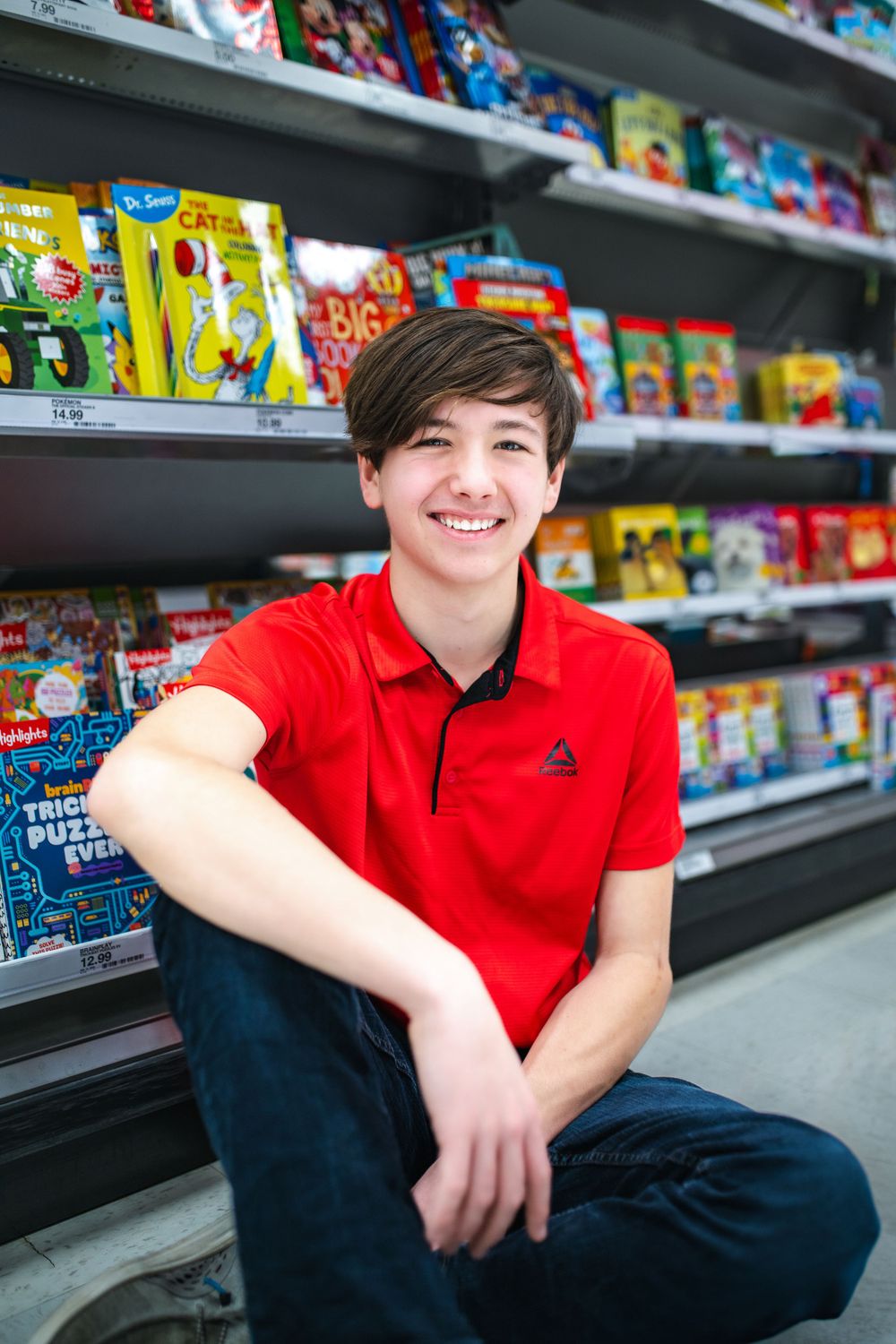A stock associate in a bright red polo shirt sits casually by shelves filled with colorful snacks and food products.