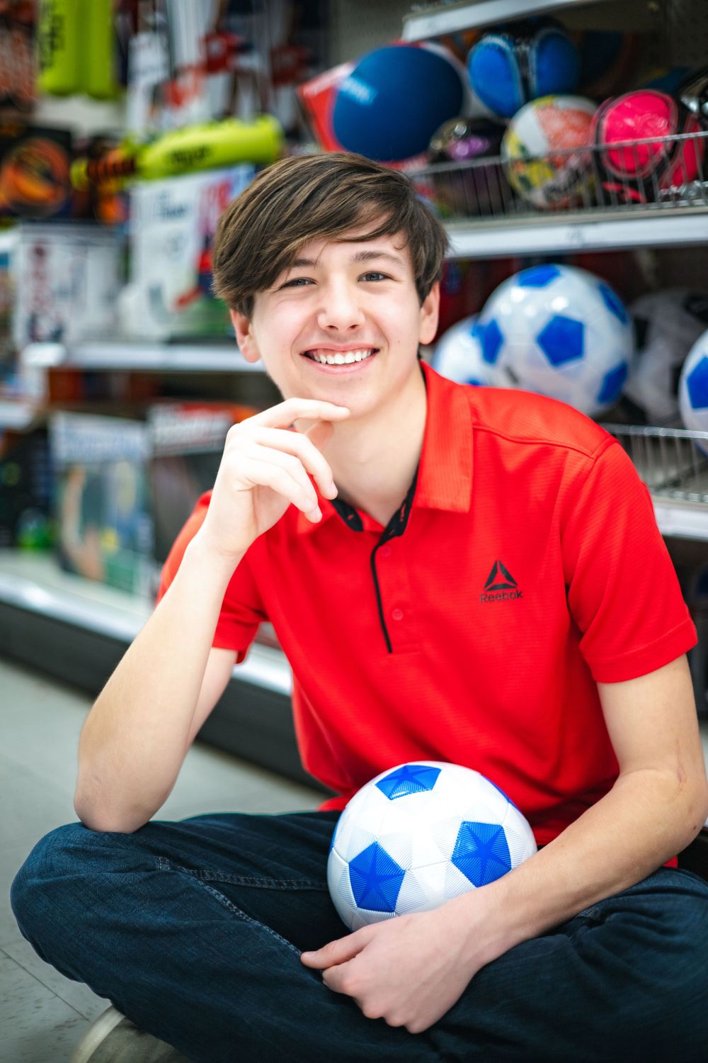 A person in a red polo shirt sits on the floor holding a blue and white soccer ball in a sporting goods store.