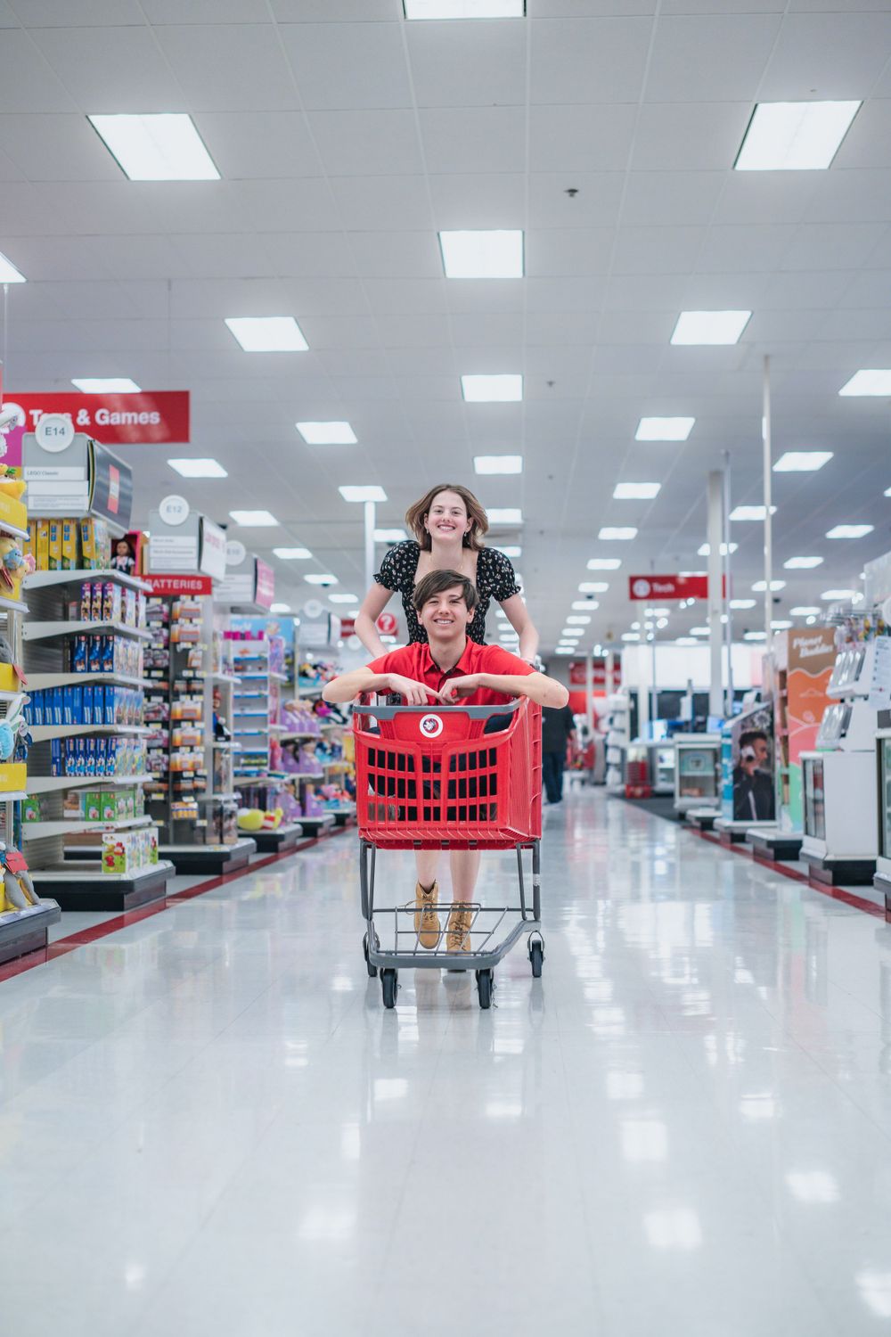 Two people pushing a red shopping cart down an empty aisle in a well-lit retail store with fluorescent lighting overhead.