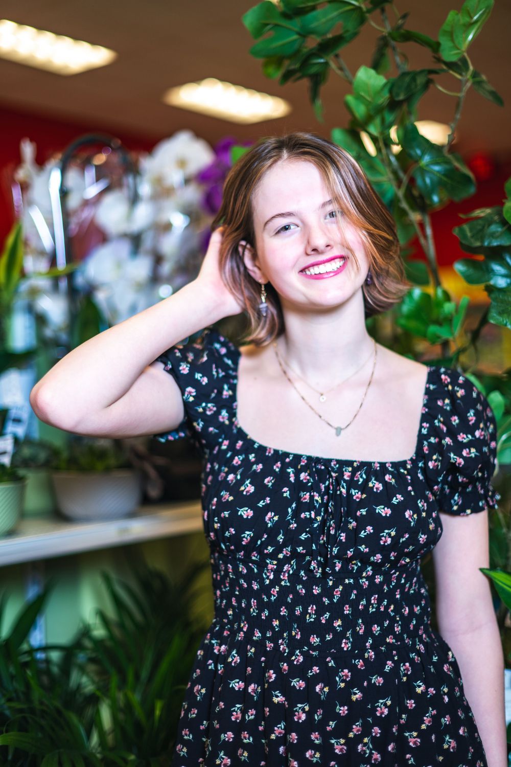 A smiling figure in a floral print dress poses near indoor plants in a room with fluorescent lighting.