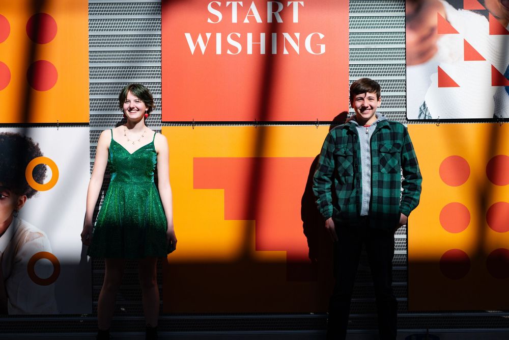 Two people stand against a vibrant orange wall with text 'START WISHING', one in green dress and one in plaid coat.