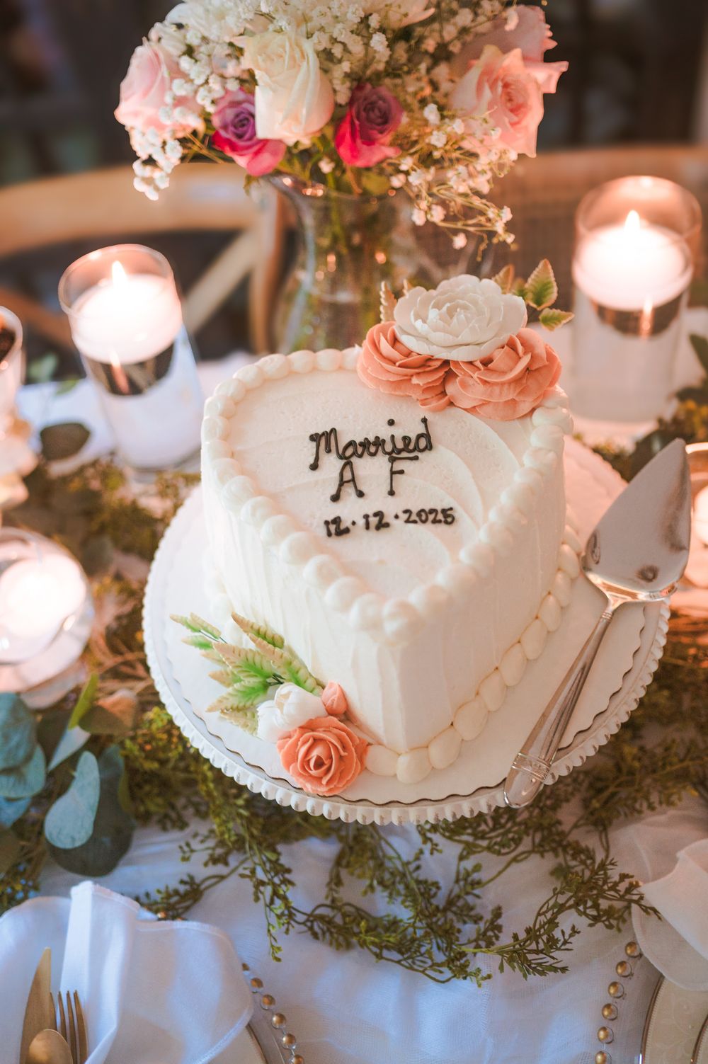 Elegant white wedding cake decorated with coral roses and greenery on a candlelit table.