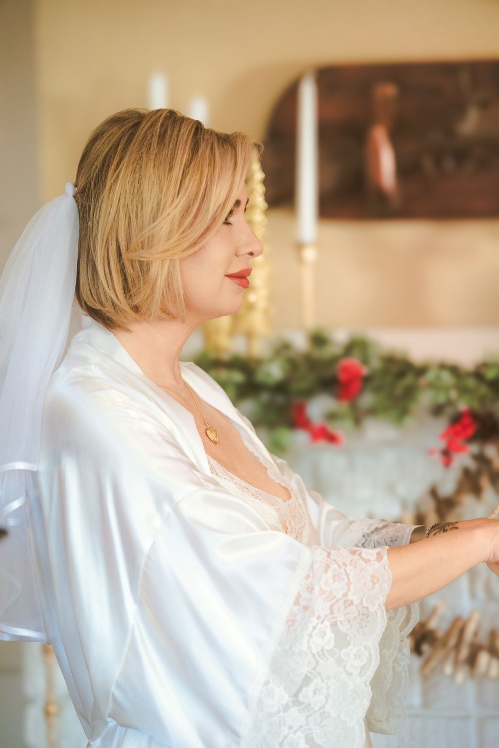 A bride in a white lace robe poses near candlelit decorations and red flowers in a romantic setting.