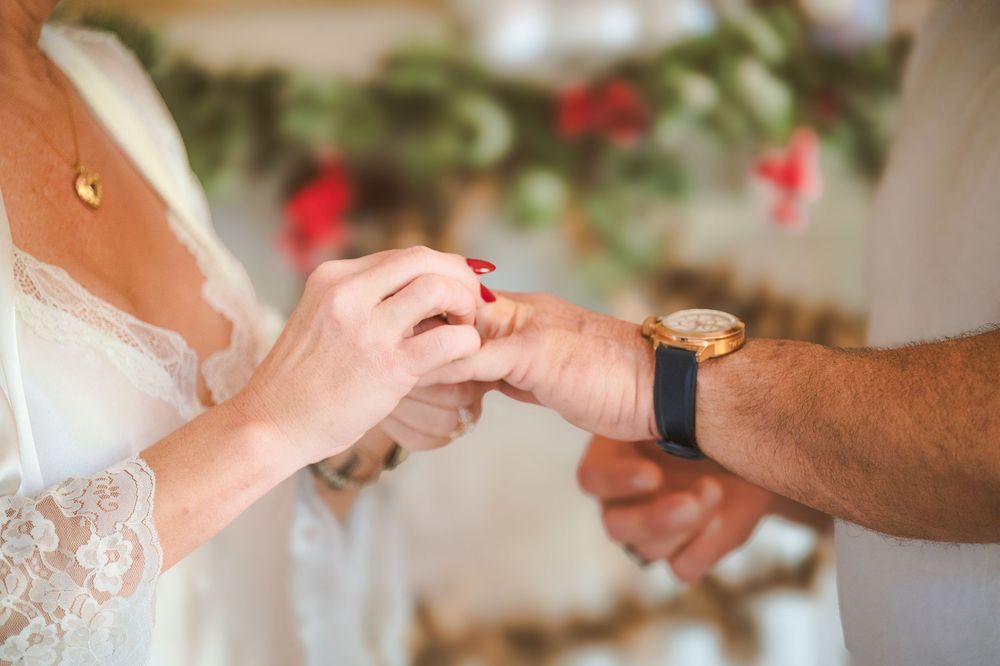Close-up of hands exchanging wedding rings during a romantic outdoor ceremony with red flowers in background.