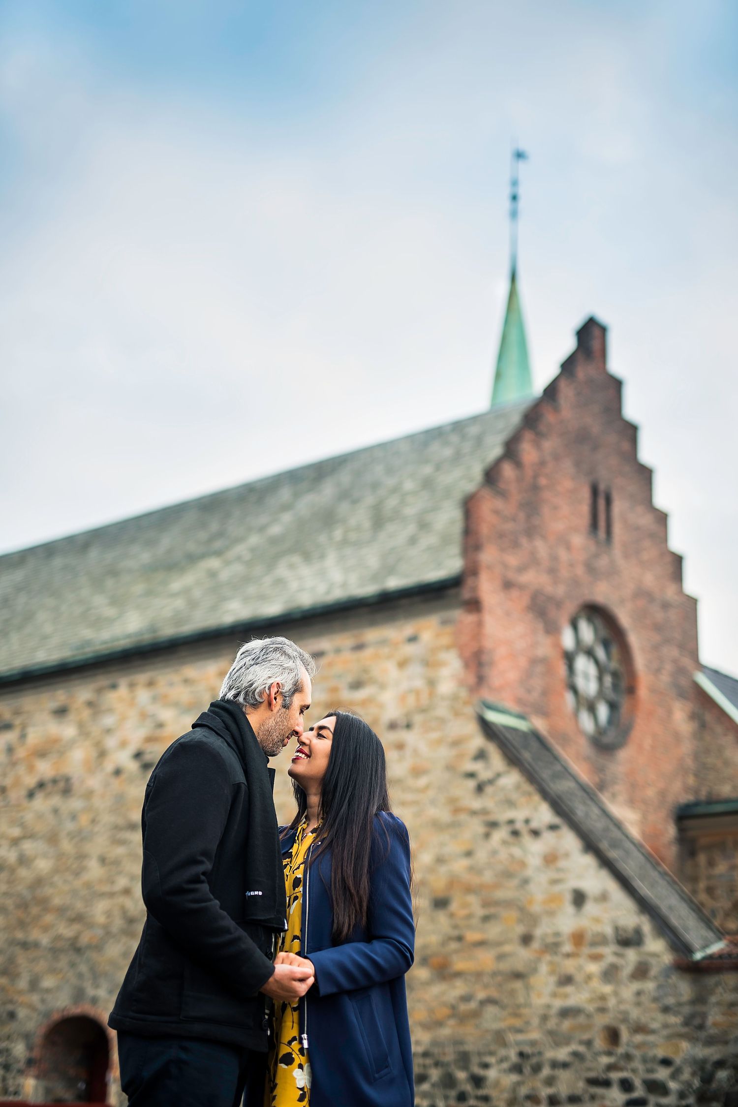 A romantic couple shares an intimate moment while standing outside a historic stone church with a spired roof.