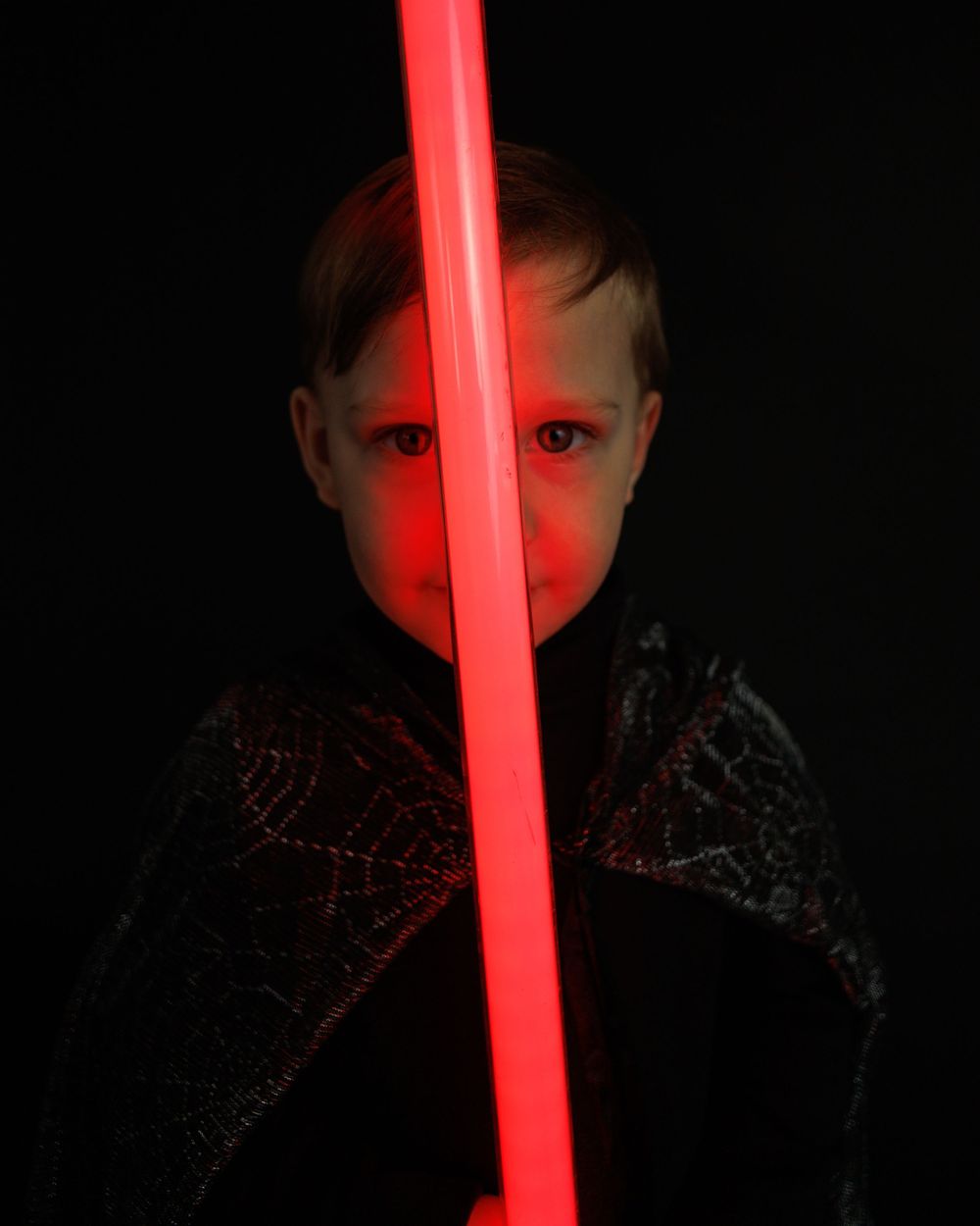 Figure illuminated by red glow from lightsaber prop against dark background in dramatic portrait.