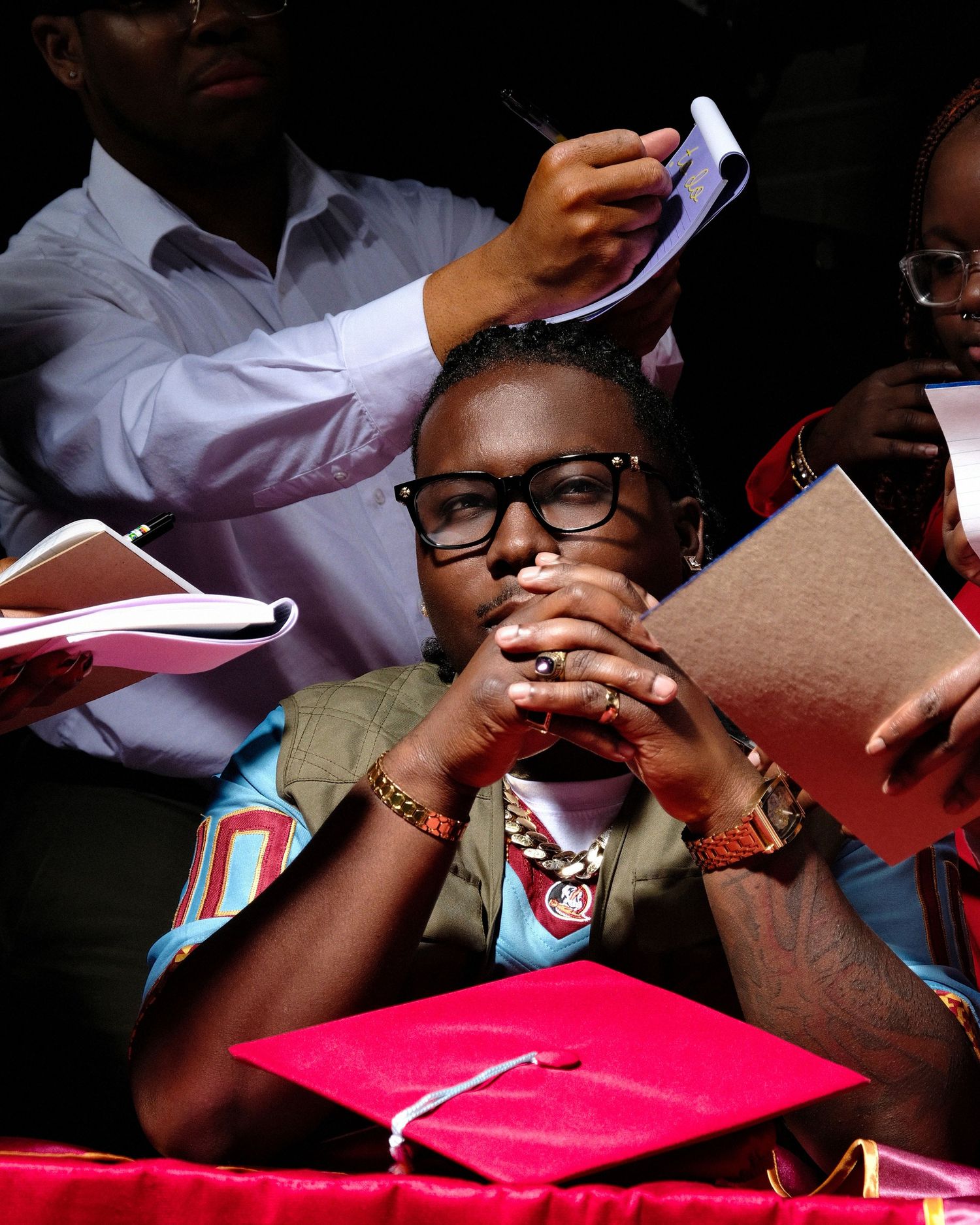 Student in glasses amid graduation ceremony wearing olive green attire, with a bright pink diploma cover visible.