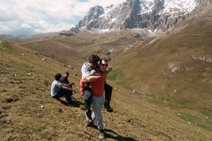 Group of hikers resting on a grassy mountain slope with dramatic peaks in the distance.
