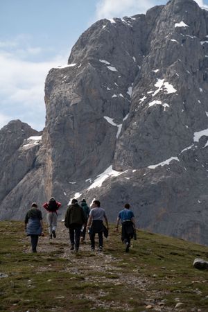 Hikers ascending a grassy slope beneath towering rocky mountain cliffs.